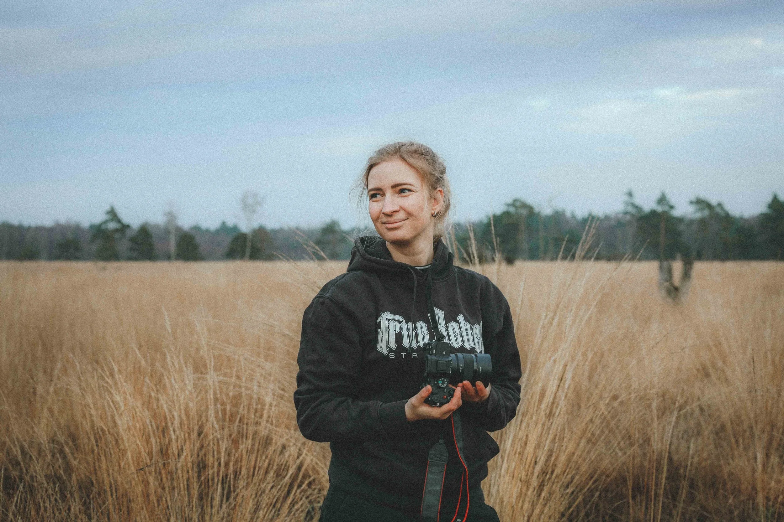 Eine junge Frau mit einem schwarzen Hoodie, die in einem Getreidefeld steht und eine Kamera in der Hand hält, bei bewölktem Himmel.