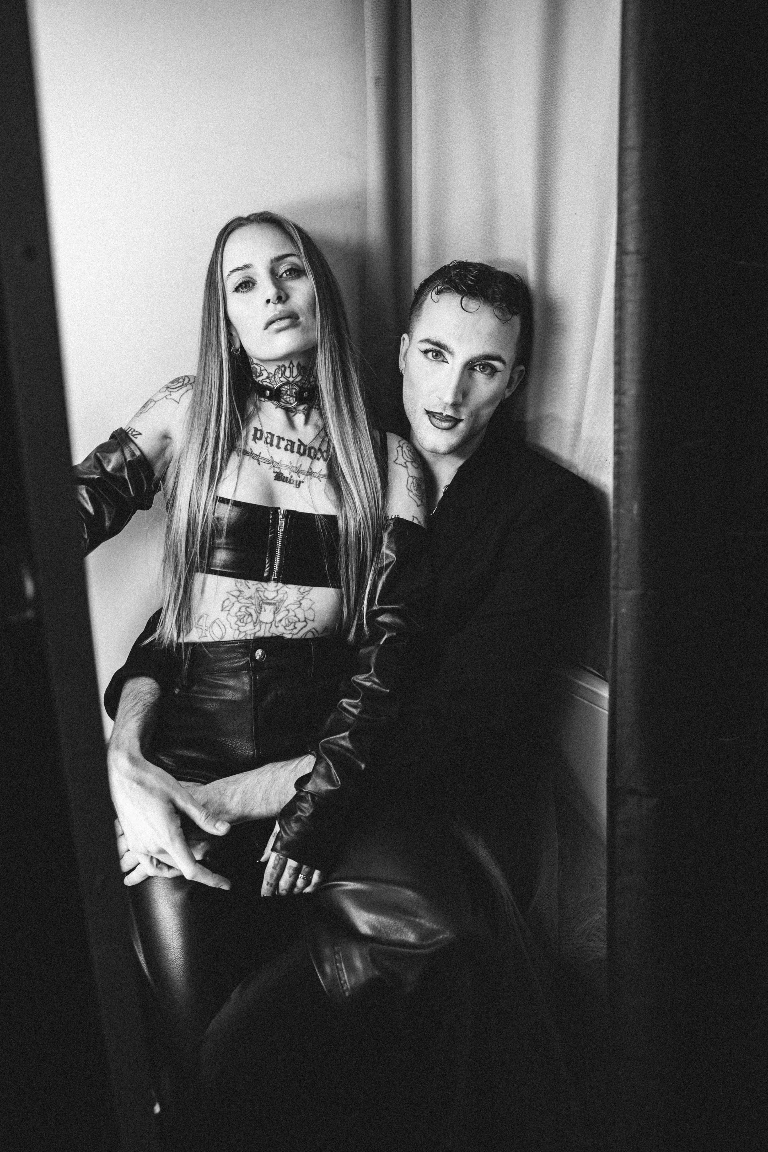 Zwei tätowierte Personen in schwarzer Kleidung posieren im Schwarzweiß-Foto, eine sitzt und eine steht hinter ihr.