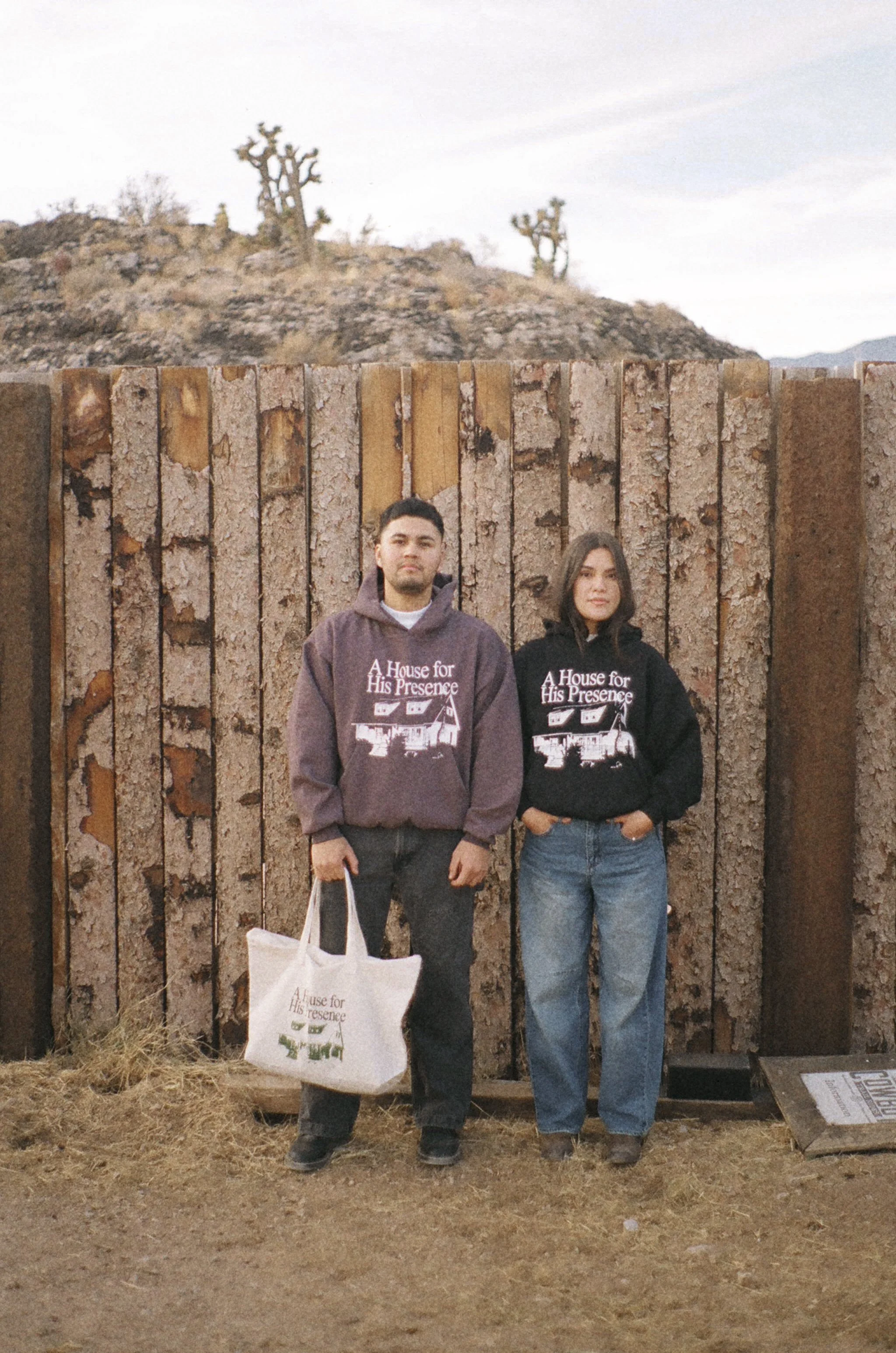 Two young adults standing in front of a wooden fence with desert landscape and Joshua trees in the background. Both are wearing hoodies with the phrase 'A House for His Presence' and an illustration of a house.