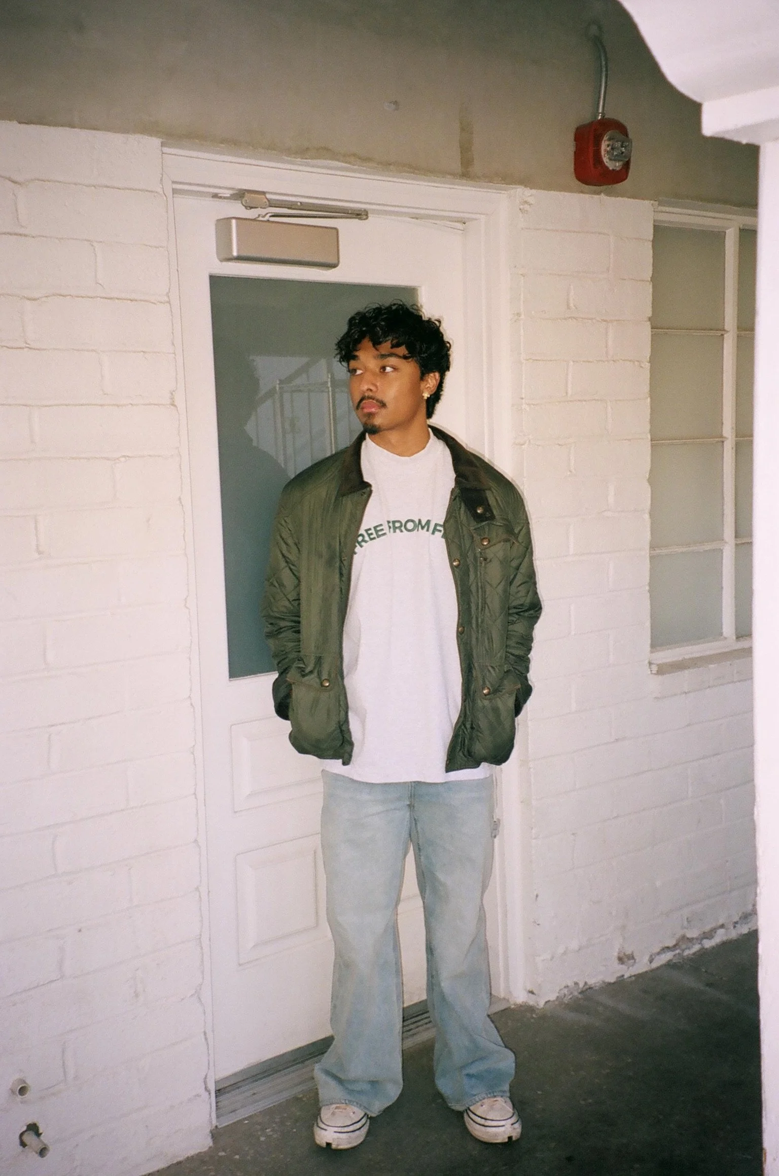 A young man standing in front of a white brick wall and a glass door, wearing a green jacket, white T-shirt, light-wash jeans, and sneakers, with hands in his pockets, looking to his left.