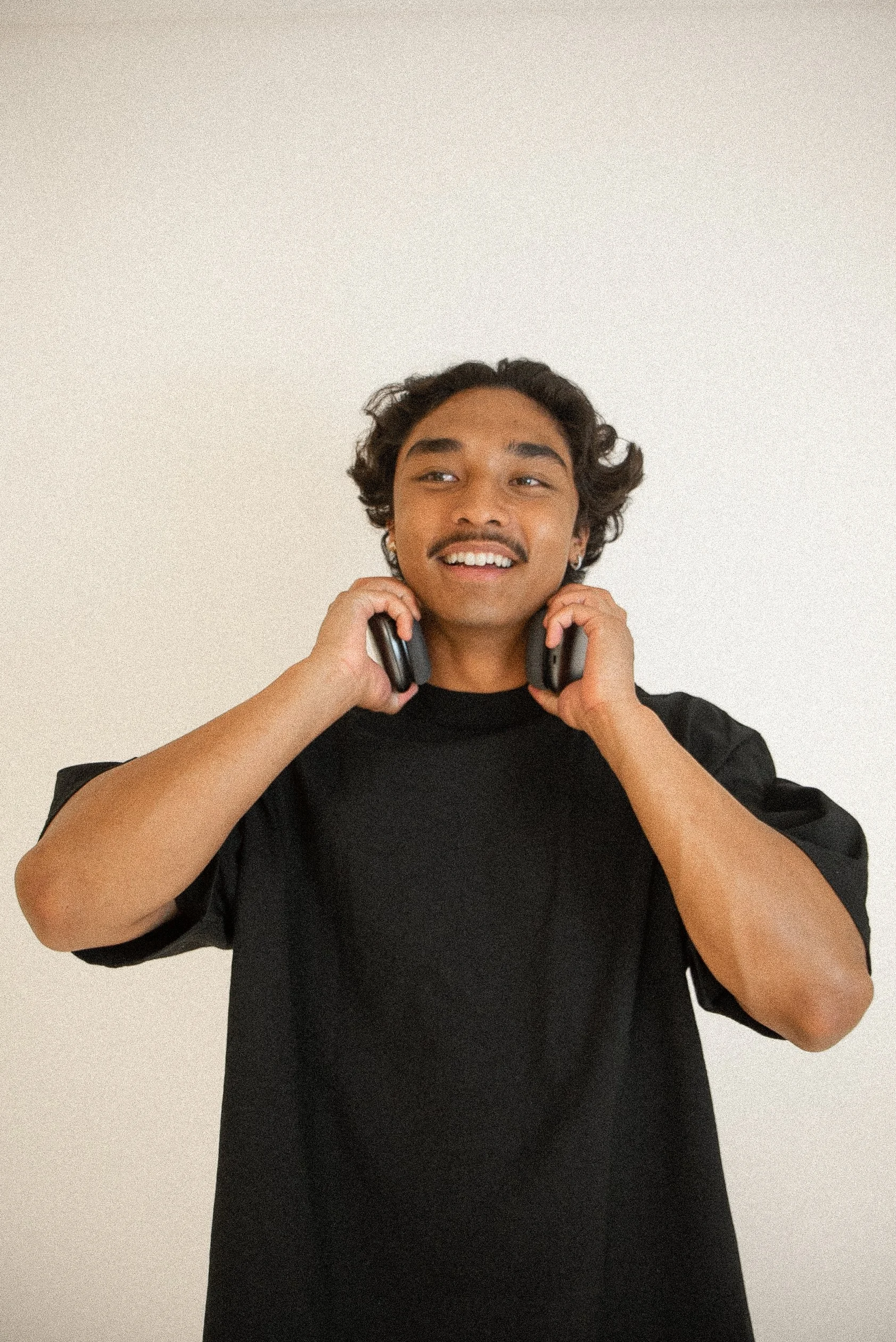 Young man with curly hair and mustache smiling, holding headphones around his neck, wearing a black t-shirt against a plain light-colored background.