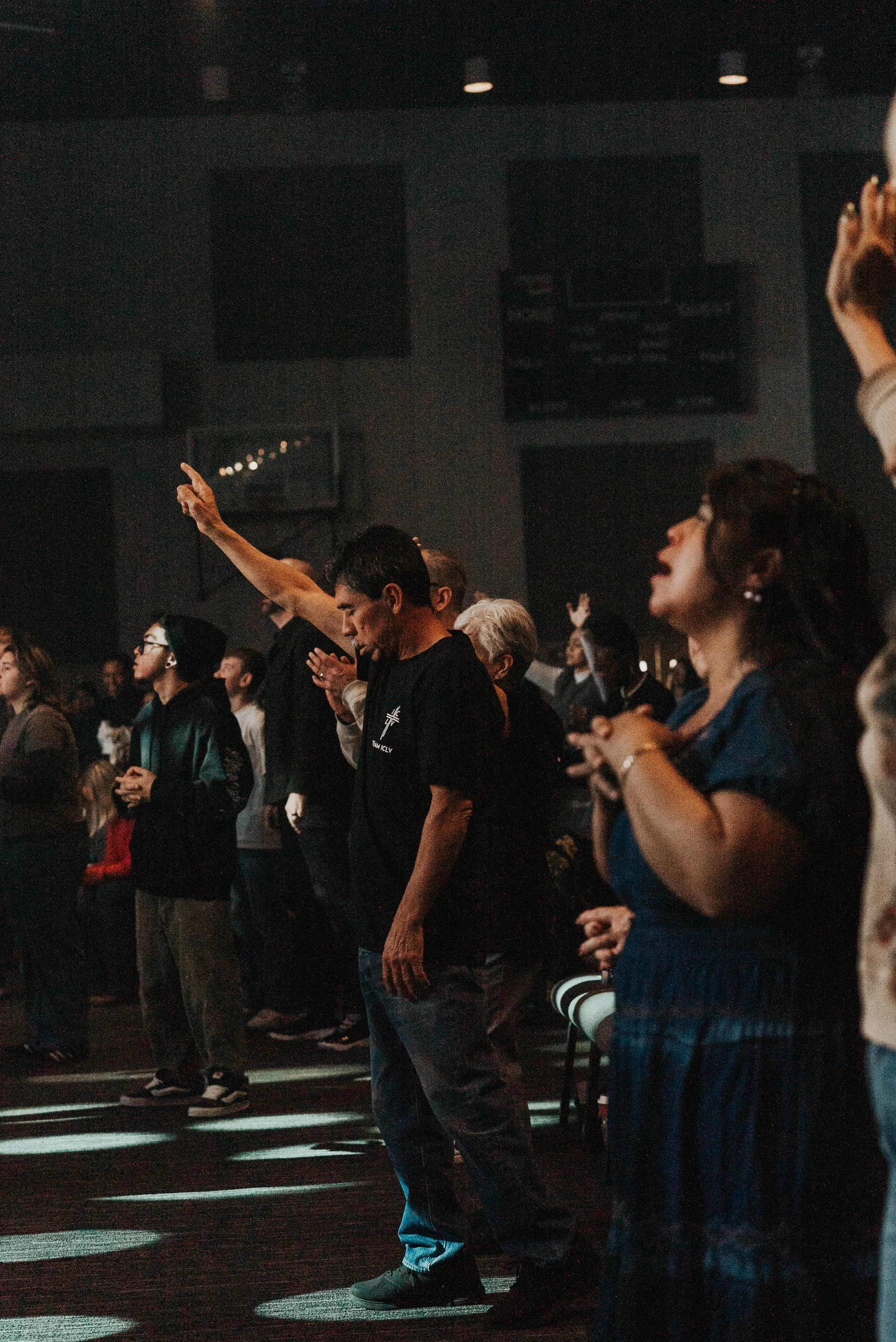 People attending a worship service, some with hands raised and eyes closed, inside a darkened auditorium.