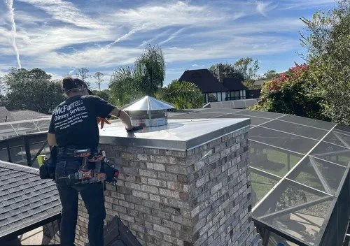A person installing a chimney cap on a rooftop with a house and trees in the background.