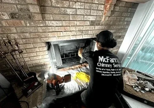 A chimney sweep worker cleaning a fireplace, wearing a black, hat, and stickers on the back of his shirt reading 'McFarran Chimney Services'.