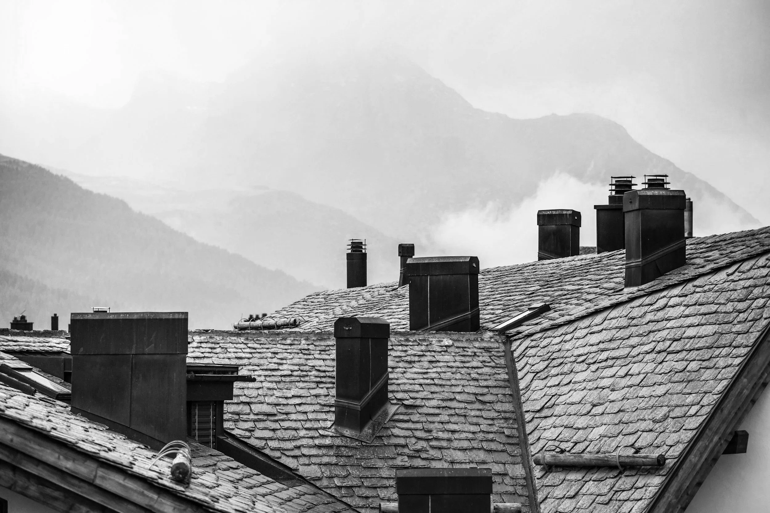 Black and white photograph of rooftops with multiple chimneys, set against mountains and misty sky in the background.