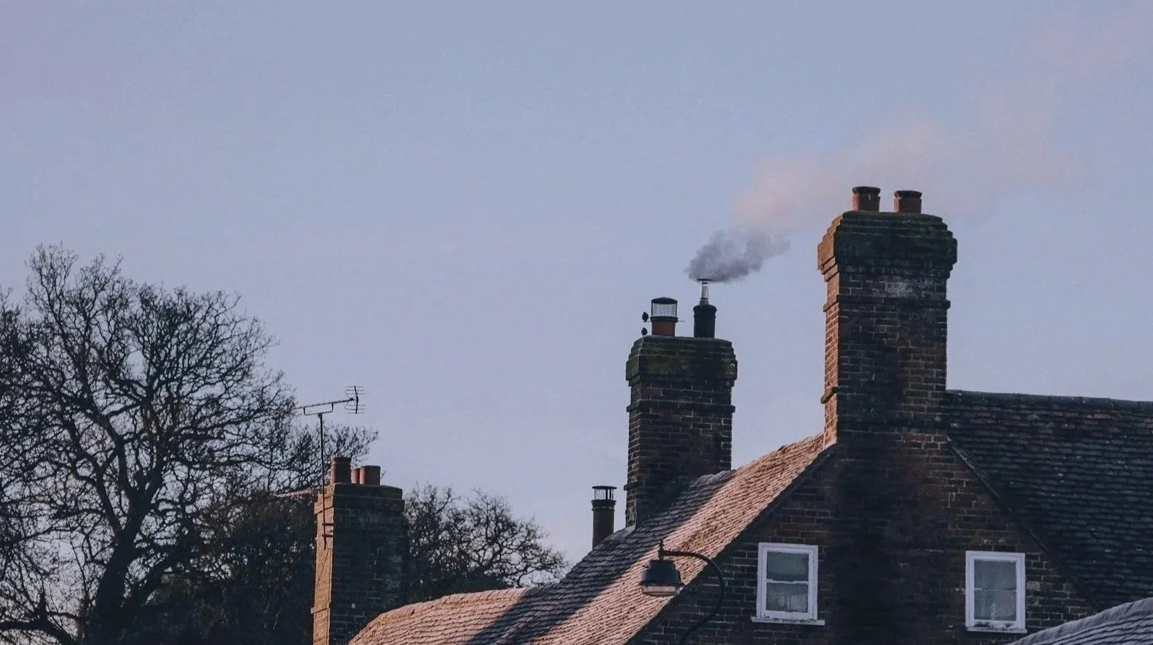 A photograph of rooftops of brick houses with multiple chimneys, one of which is emitting white smoke. There is a leafless tree and a TV antenna against a light sky.