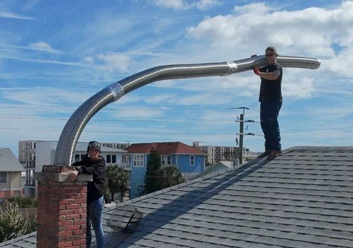 Two people installing a large metal vent pipe for a chimney on a residential roof with blue skies and clouds in the background.