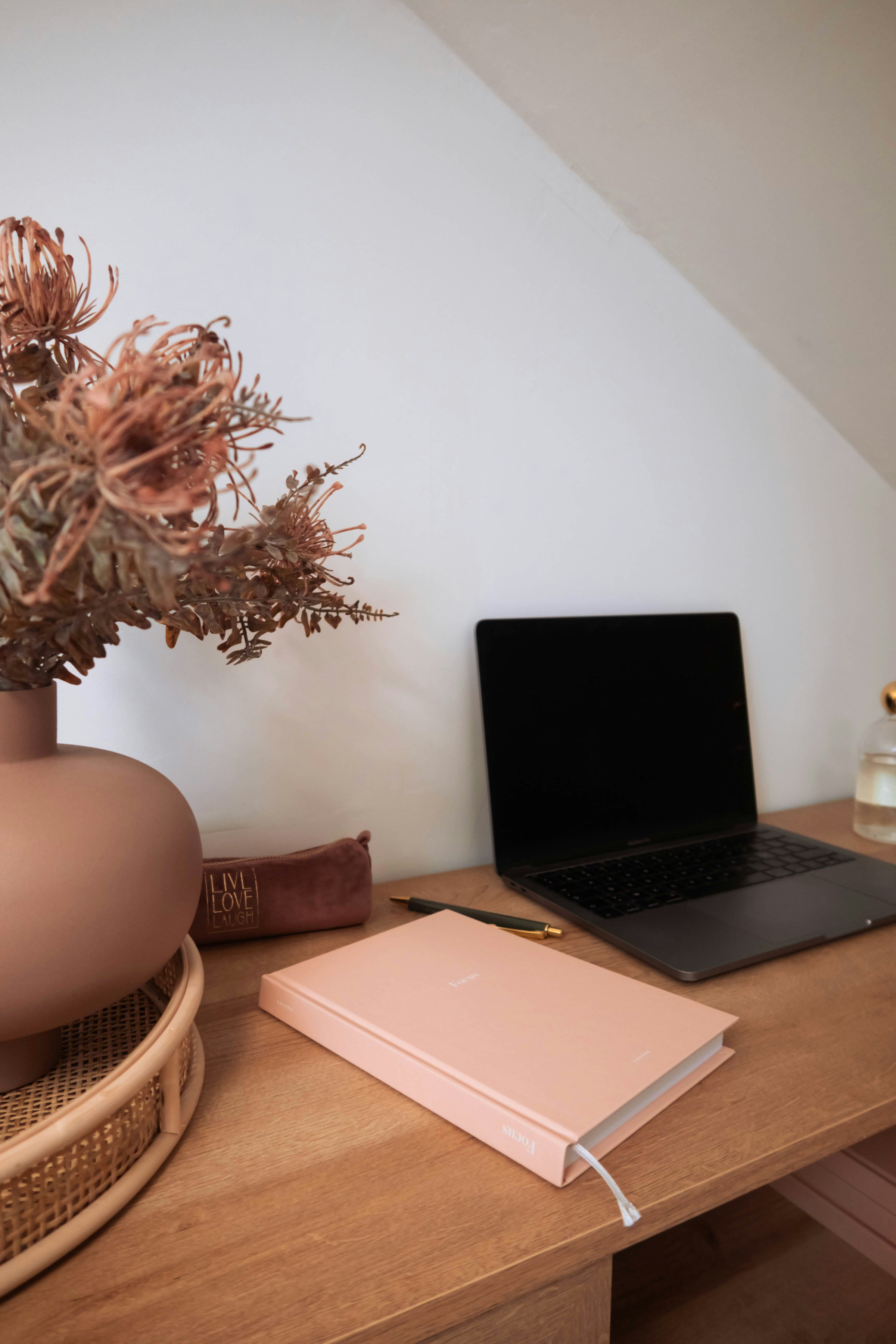 A wooden desk with a pink notebook, gold pen, closed laptop, a vase with dried flowers, and a pink pencil case with gold text, against a white wall.
