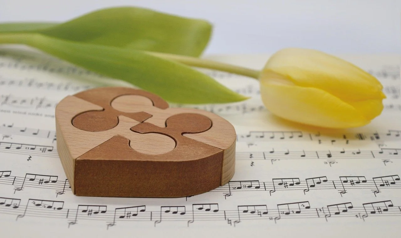 A yellow tulip flower with green leaves resting on sheet music with musical notes, and a wooden puzzle piece on top.