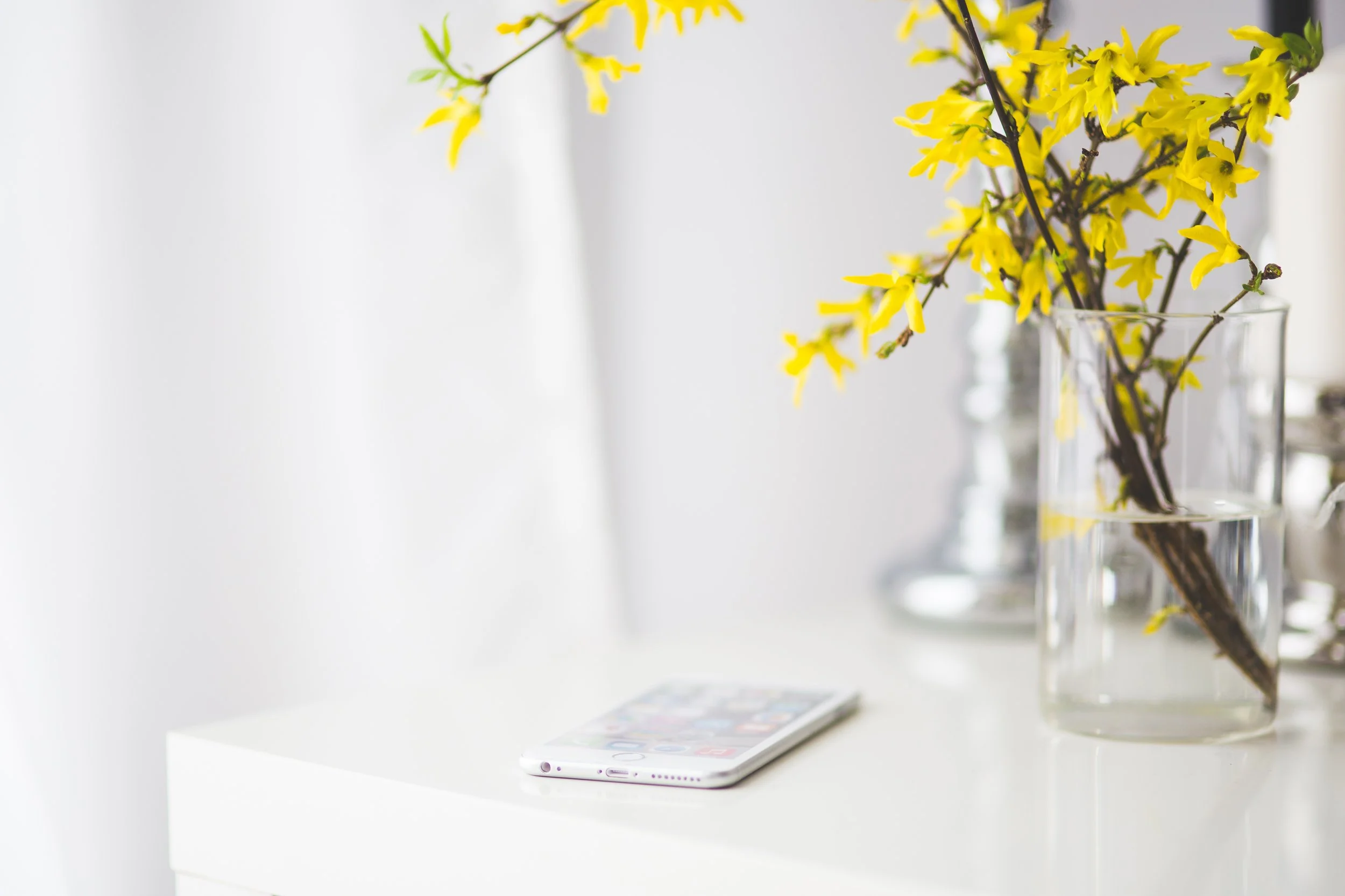 A white smartphone lying flat on a white surface with a yellow flower arrangement in a glass vase nearby.