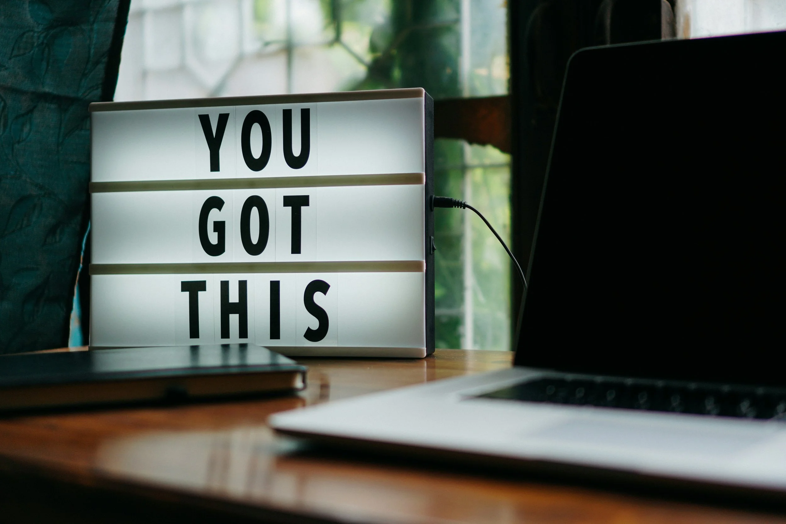 A lightbox sign with the words 'You Got This' in black letters, placed on a wooden desk next to a laptop and a closed notebook, in a room with a window in the background.