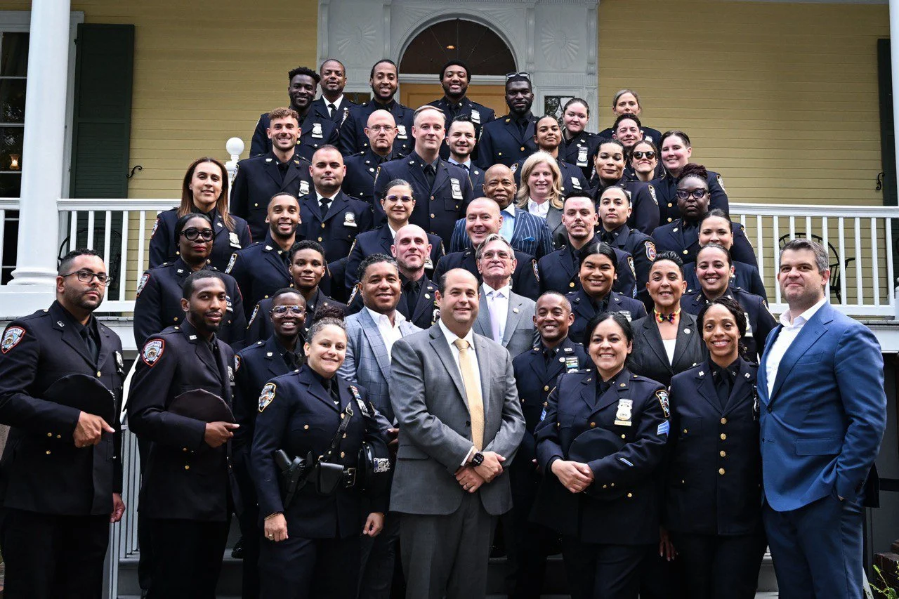 A large group of police officers and officials posing together on the steps of a yellow house with white trim. The group includes men and women all dressed in police uniforms or formal attire, smiling at the camera.