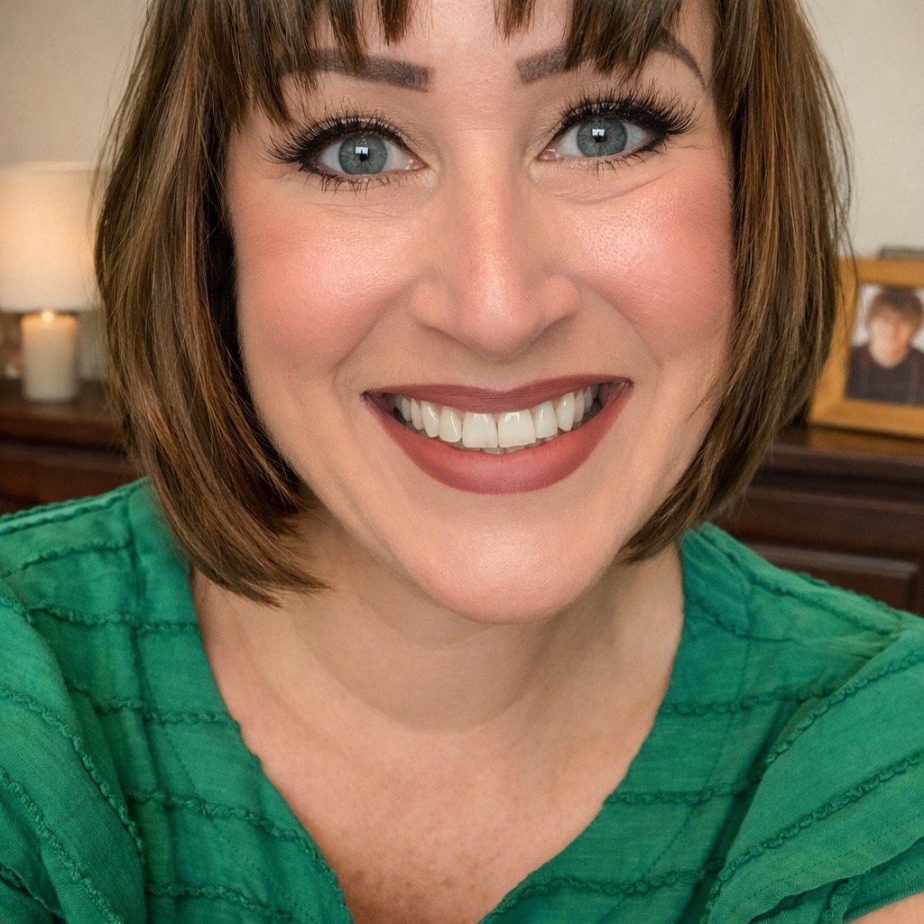 Close-up of a smiling woman with short brown hair, blue eyes, and tan skin, wearing a green top, indoors with a lamp and framed photo in the background.
