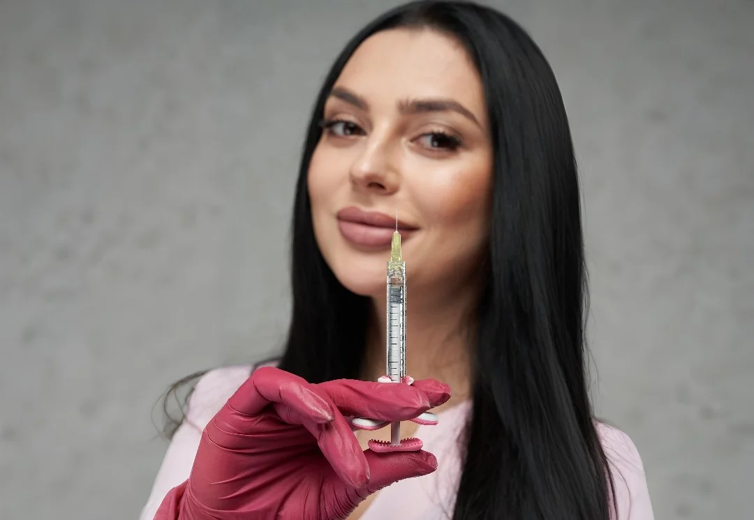 A woman with long dark hair holding a syringe and a pink glove, standing against a gray background.