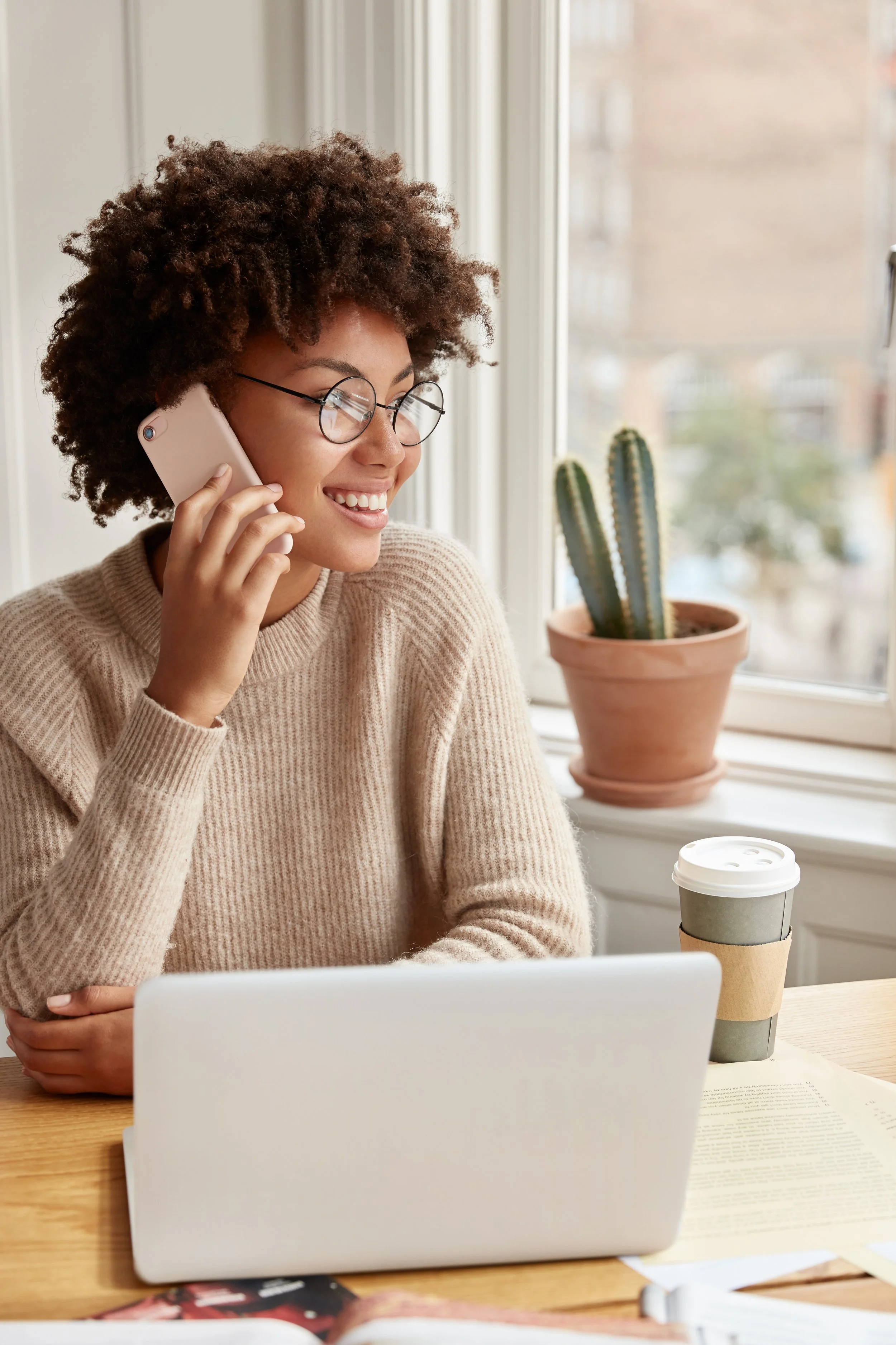 A woman with curly hair and glasses smiling while talking on a smartphone, sitting at a wooden table with a laptop and a takeaway coffee cup, next to a window with a potted cactus on the windowsill.