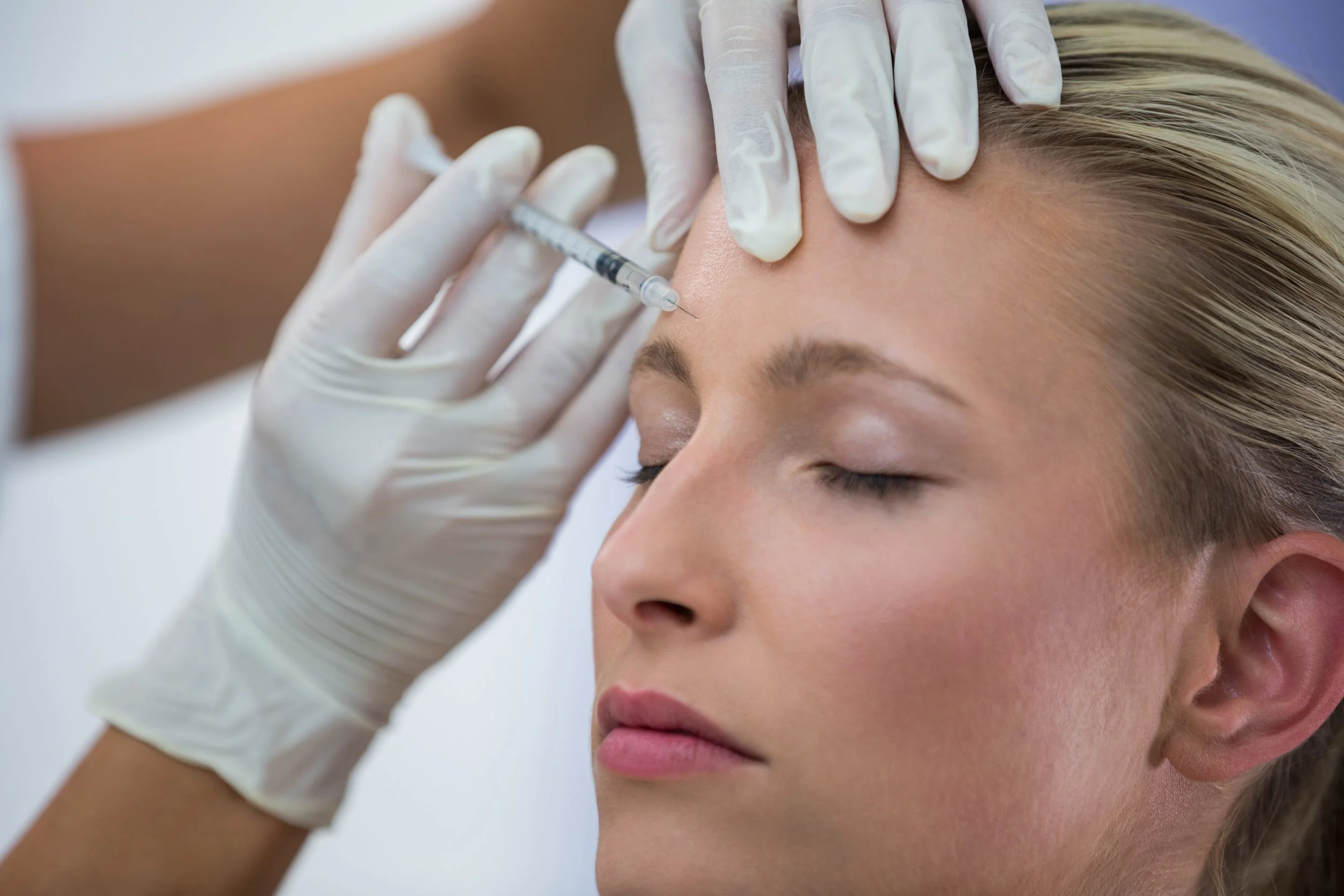 A woman receives a Botox injection in her forehead from a medical professional wearing white gloves.