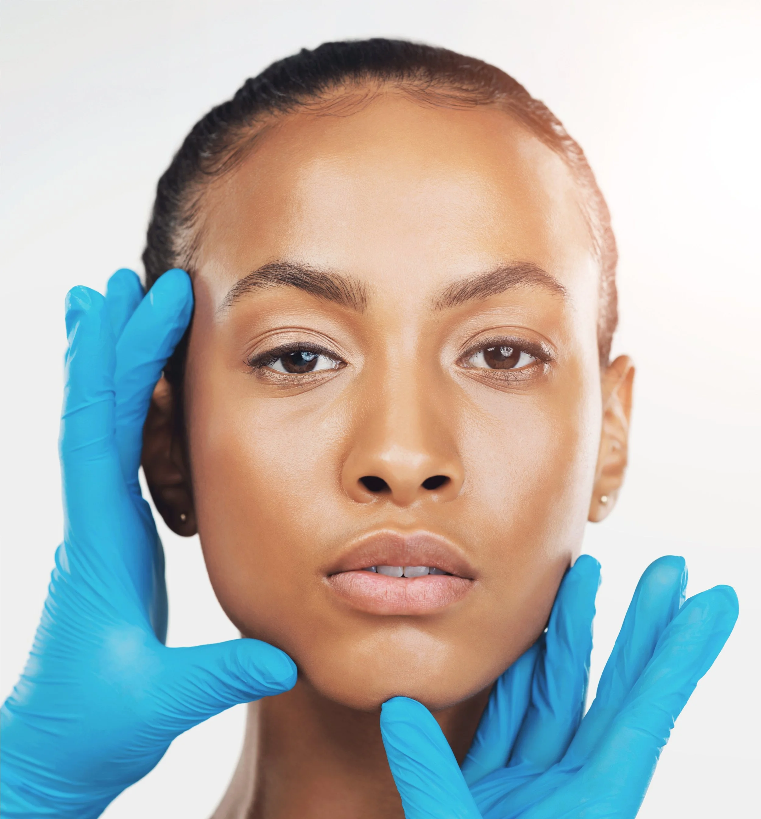 A woman with natural skin and makeup receives a facial examination from two hands wearing blue medical gloves.
