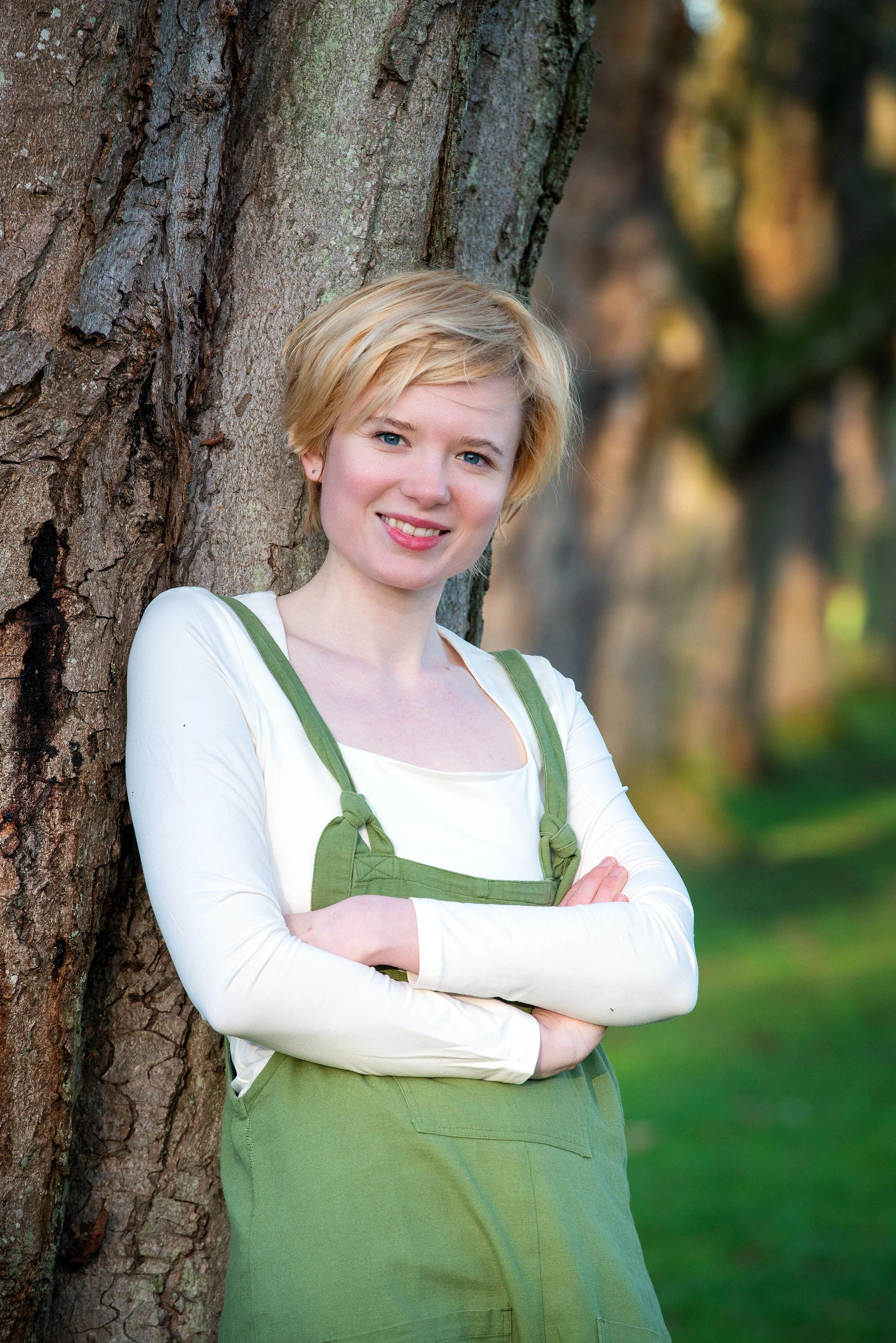Lucy Grabe-Watson, A young woman with short blonde hair and blue eyes leans against a tree outdoors, smiling at the camera. She is wearing a white long-sleeve shirt and green overalls, with arms crossed.