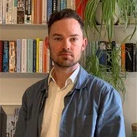 A man with short brown hair and a beard stands in front of a bookshelf filled with books, wearing a blue jacket over a beige shirt.