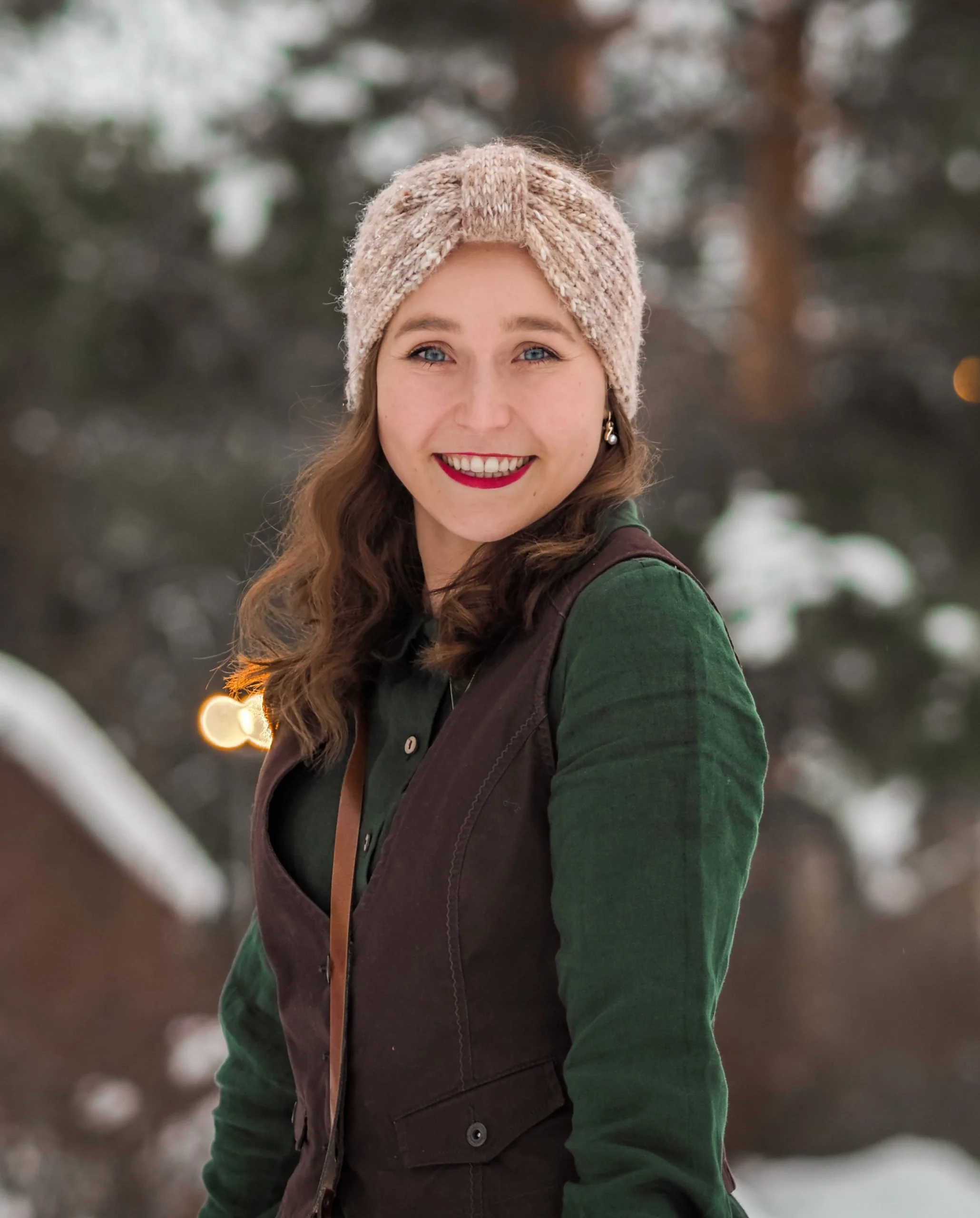 A smiling woman with blue eyes and long brown hair, wearing a light pink knit hat and green outfit, standing outdoors in snow with trees in the background.