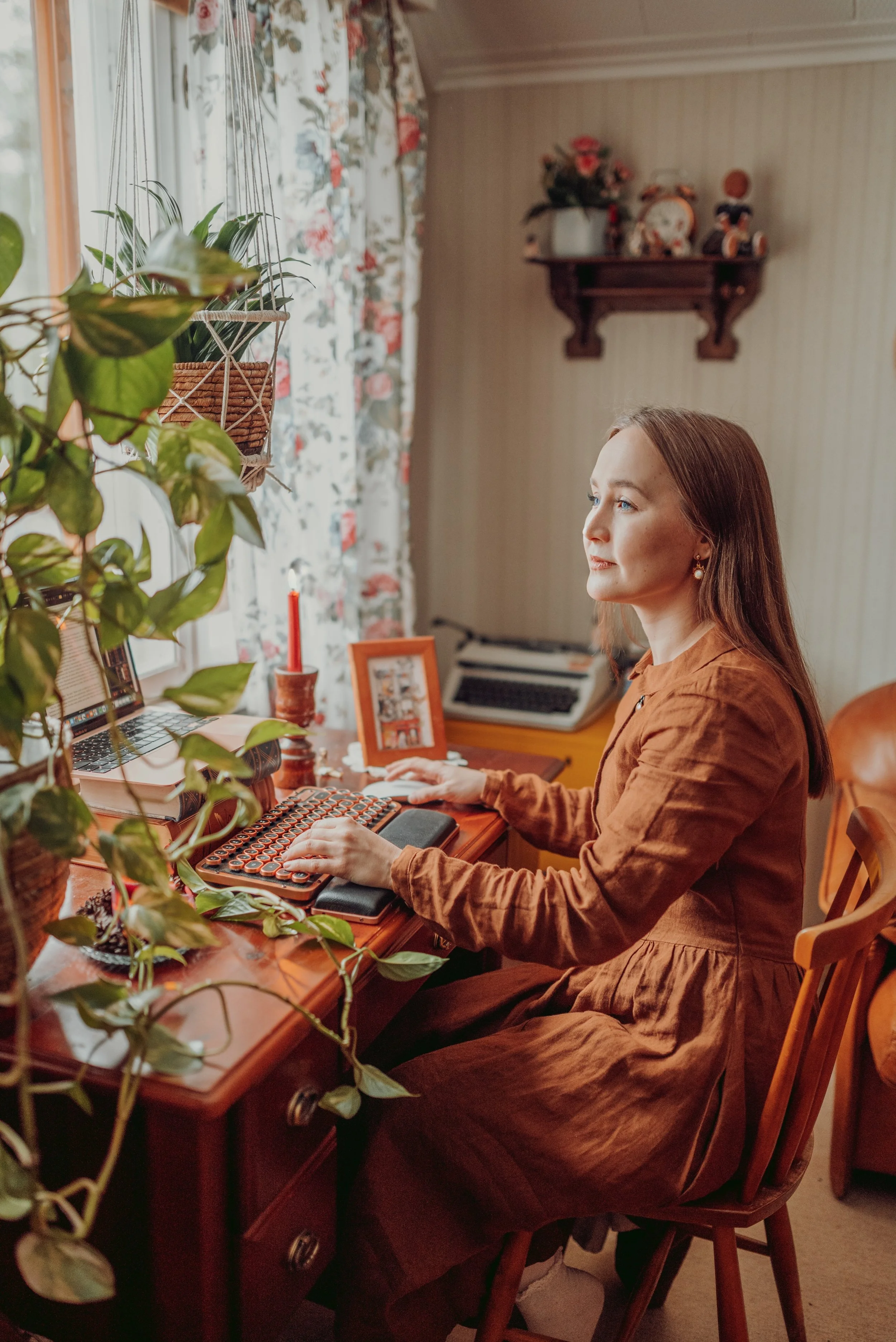 An author in a brown dress sitting at a wooden desk, working on a vintage typewriter with a laptop and decorative plants around her, in a cozy room with floral curtains and sentimental decorations.