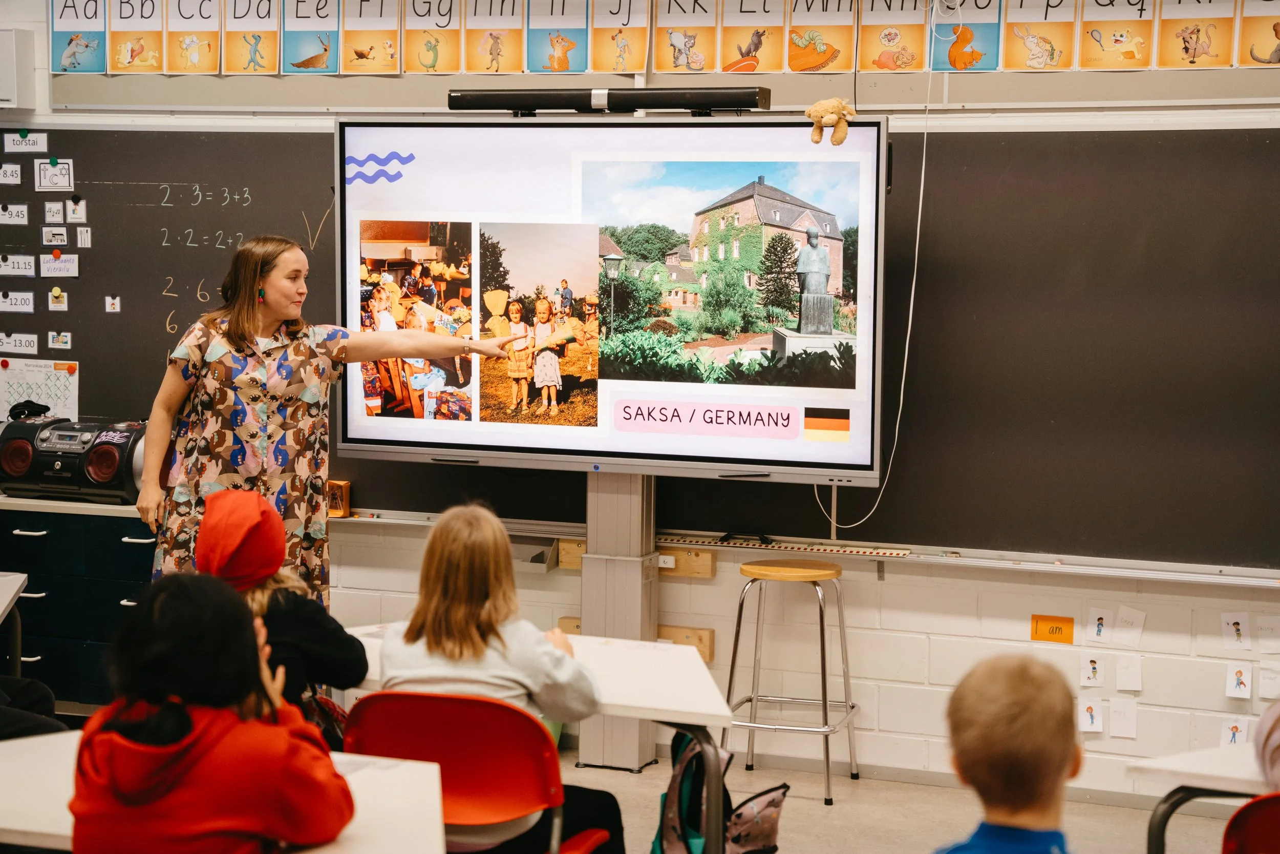 An author on a school visit points at a presentation on a large screen in a classroom, showing photos and information about her upbringing Germany. Students sit at desks facing the screen.