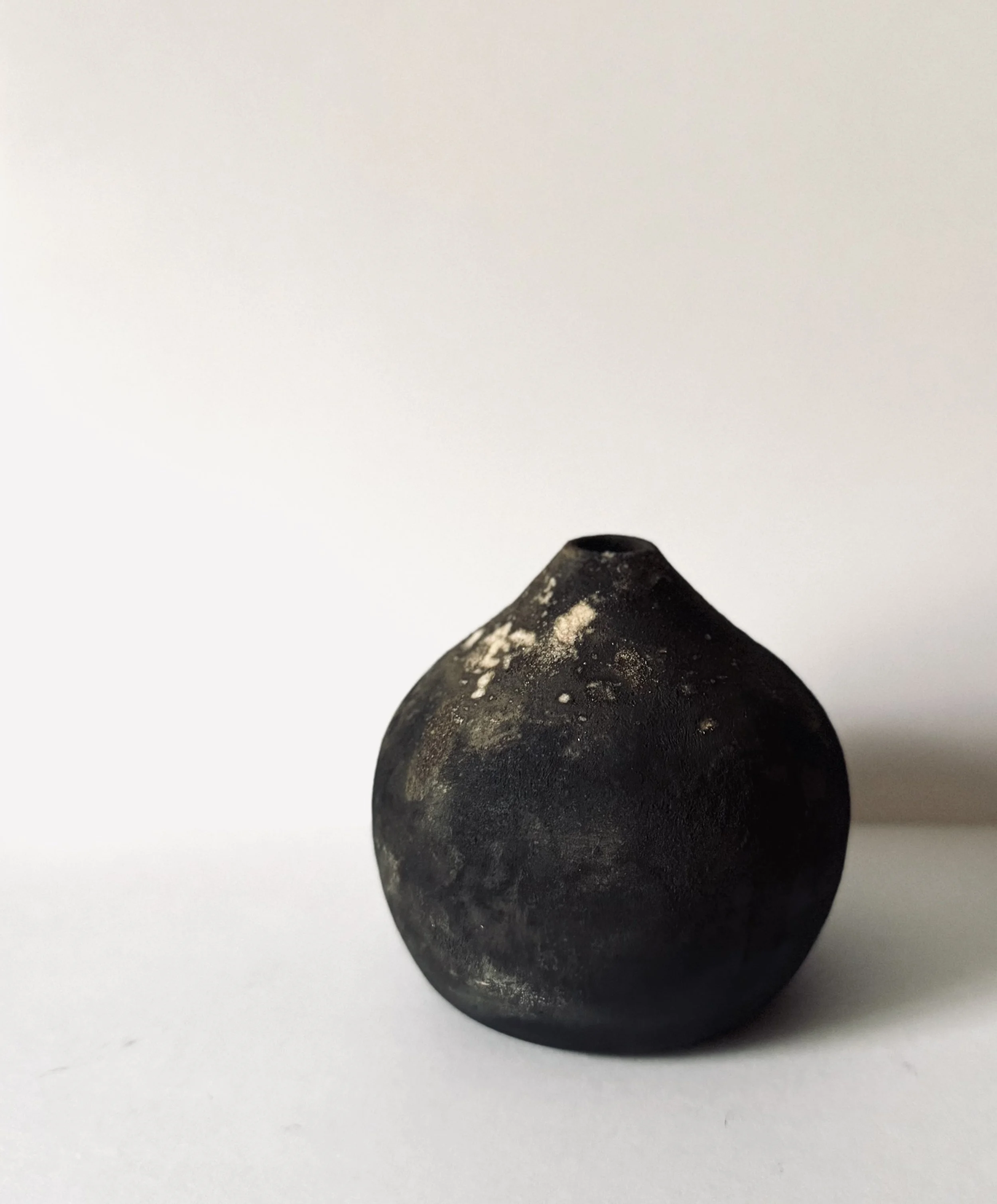 A black ceramic vase with a matte finish, slightly dusty or weathered, placed on a white surface against a plain white background.
