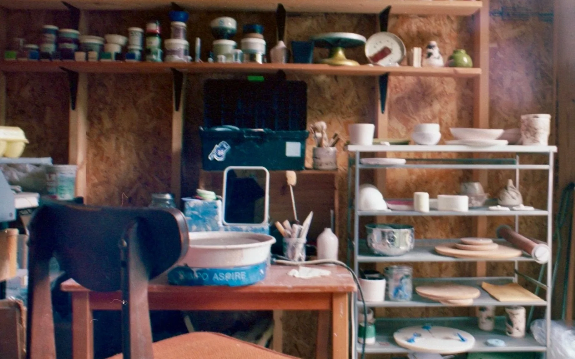 A pottery studio with shelves filled with ceramic bowls, plates, and decorative items. There is a work table with pottery tools, a small mirror, and a pottery wheel, indicating an active workspace for creating ceramics.