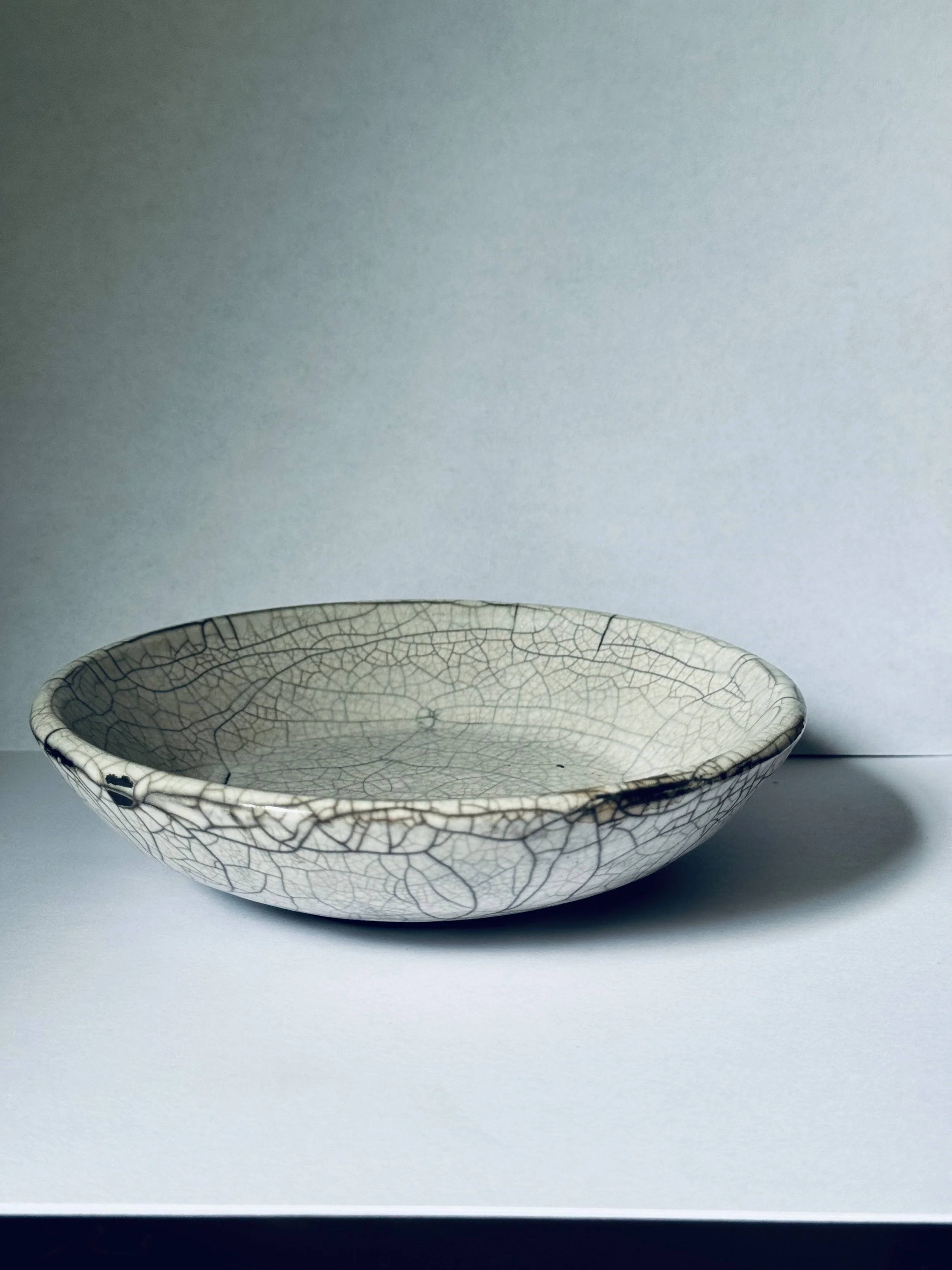 A cracked glaze ceramic bowl with a light color and intricate crackle pattern, placed on a white surface against a plain background.
