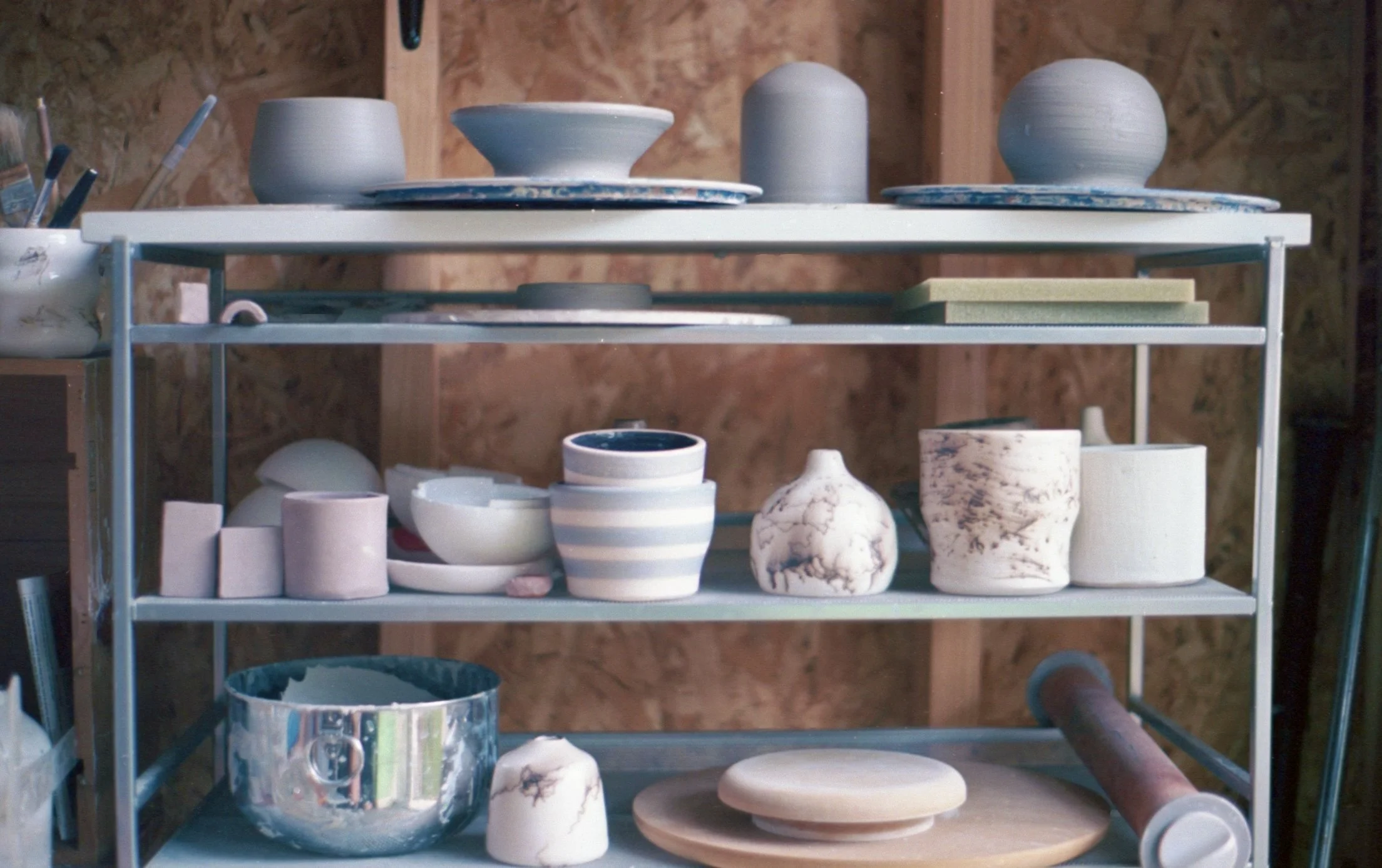 A metal shelving unit with ceramic pottery and art supplies inside a workshop with wood walls.