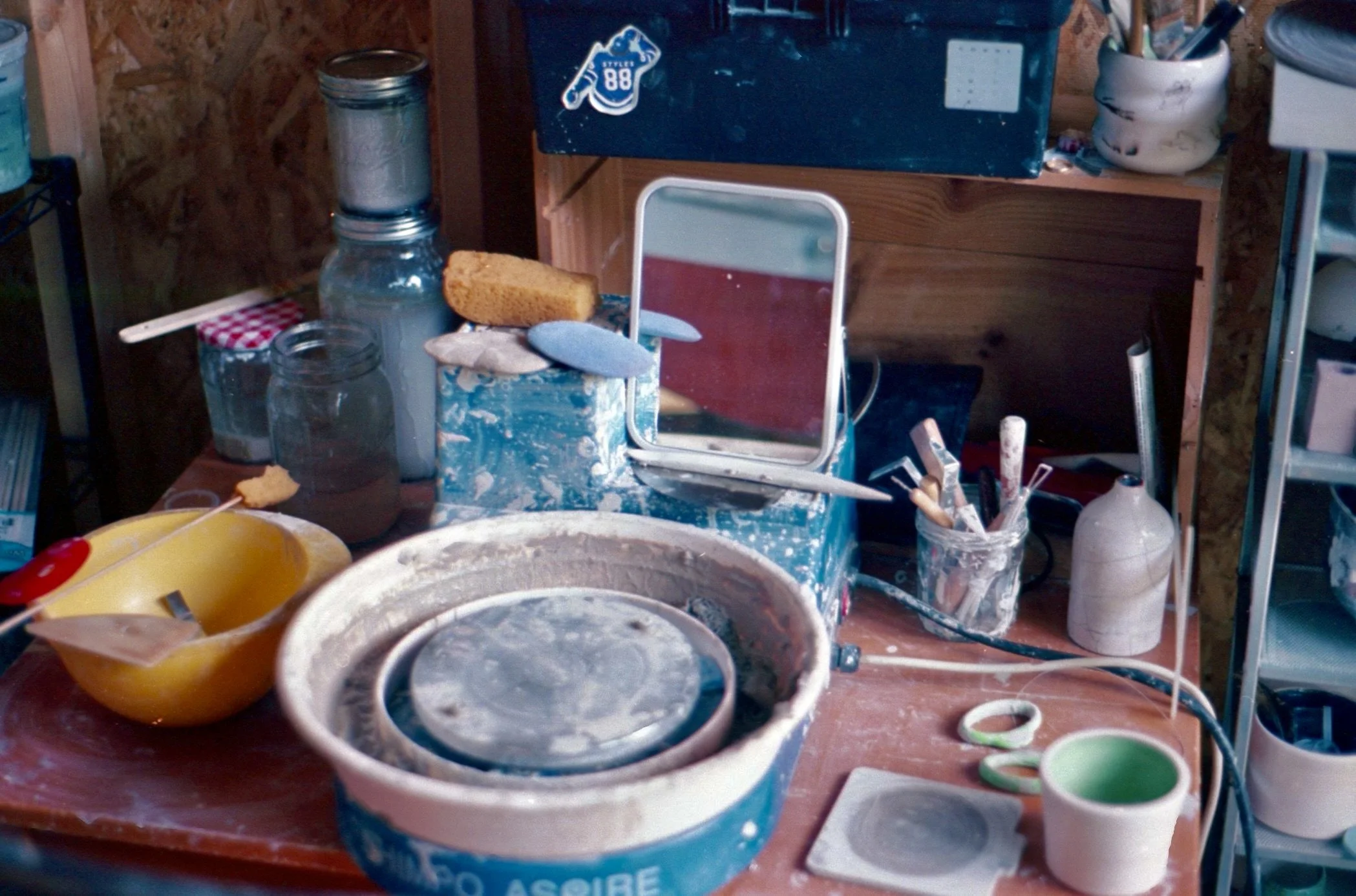 Cluttered pottery studio workspace with pottery wheel, brushes, jars, sponge, mirror, and various tools and supplies.