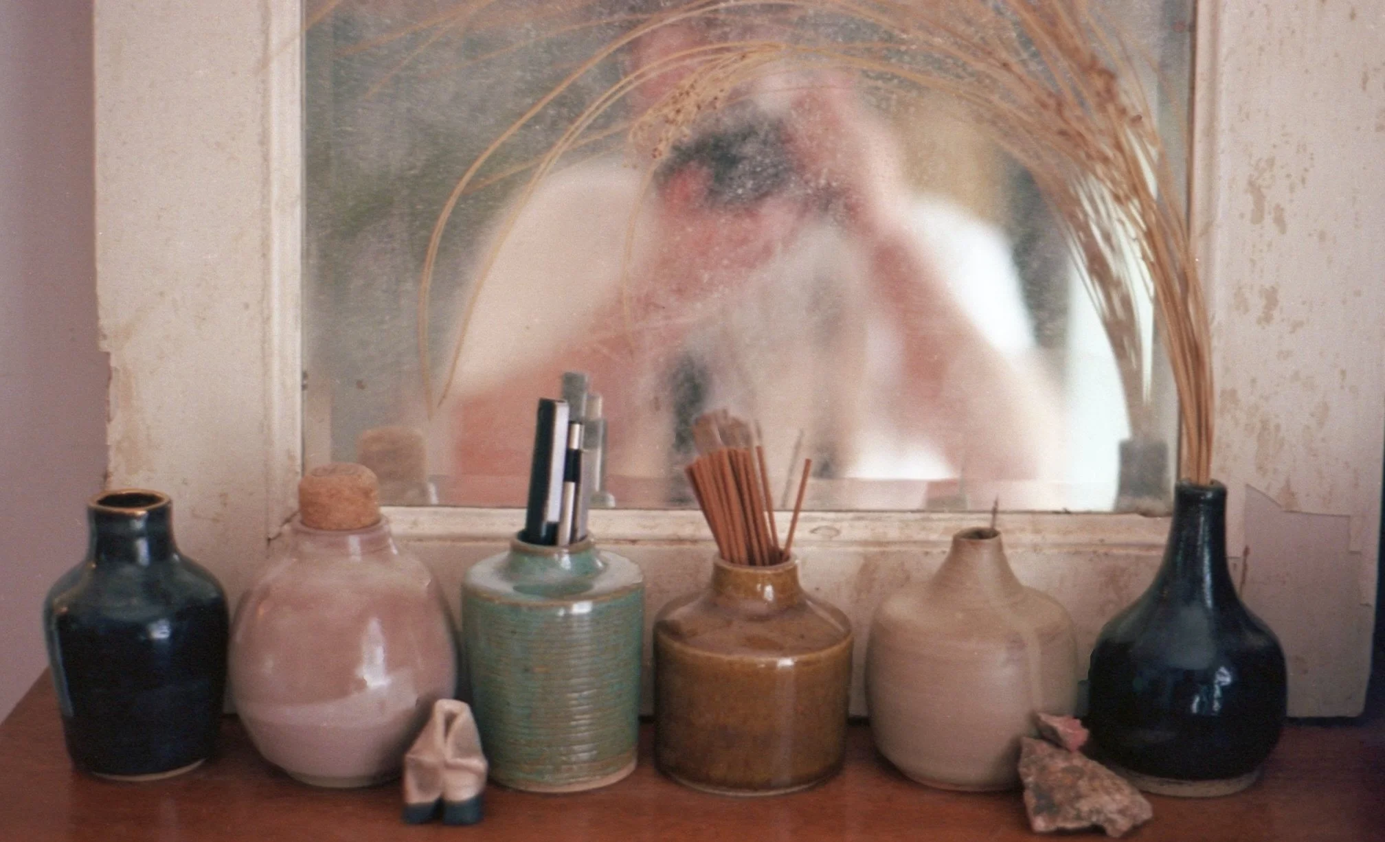 A person taking a photo of themselves in a mirror, with various ceramic vases and bottles on a wooden dresser in front of the mirror.