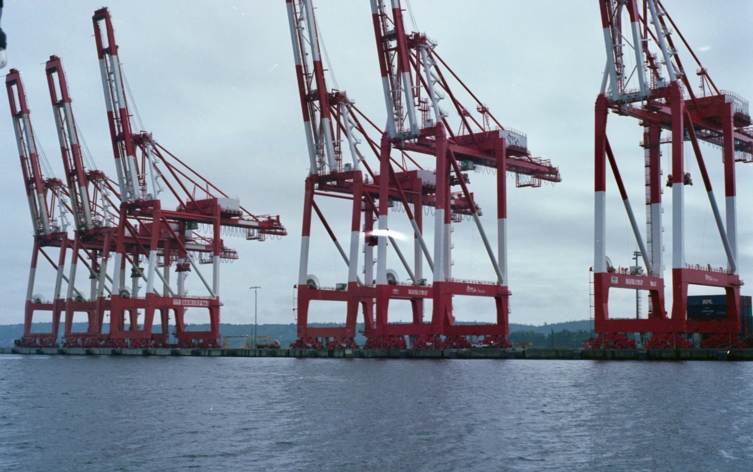 Several large red and white port cranes on a dock over water with an overcast sky.