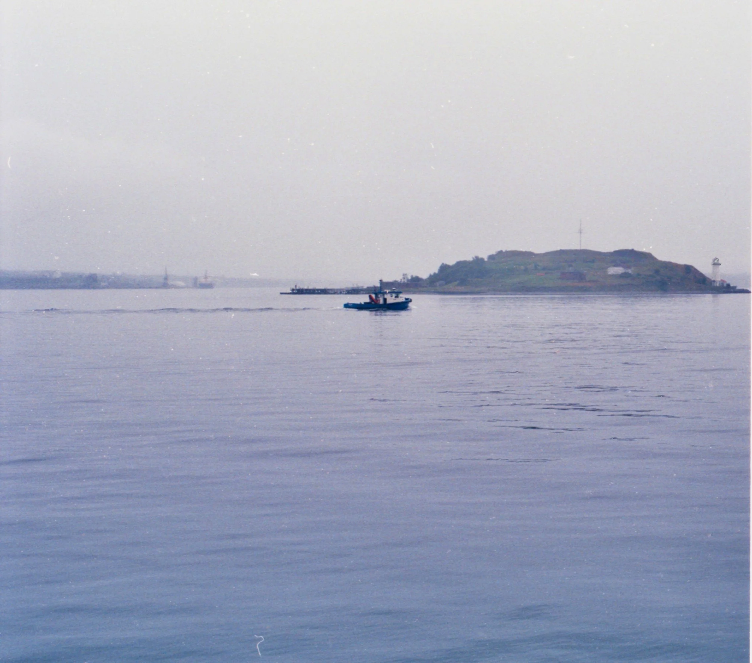 A small boat sailing on calm water near a distant hilly island with a few structures and a lighthouse, under an overcast sky.