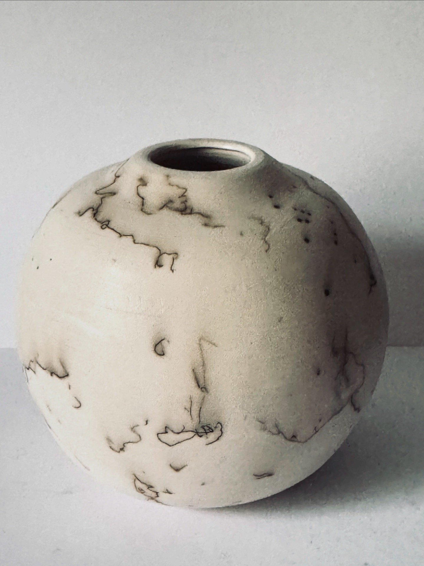 Round white ceramic vase with black marble-like veins and a small opening at the top, placed against a plain background.