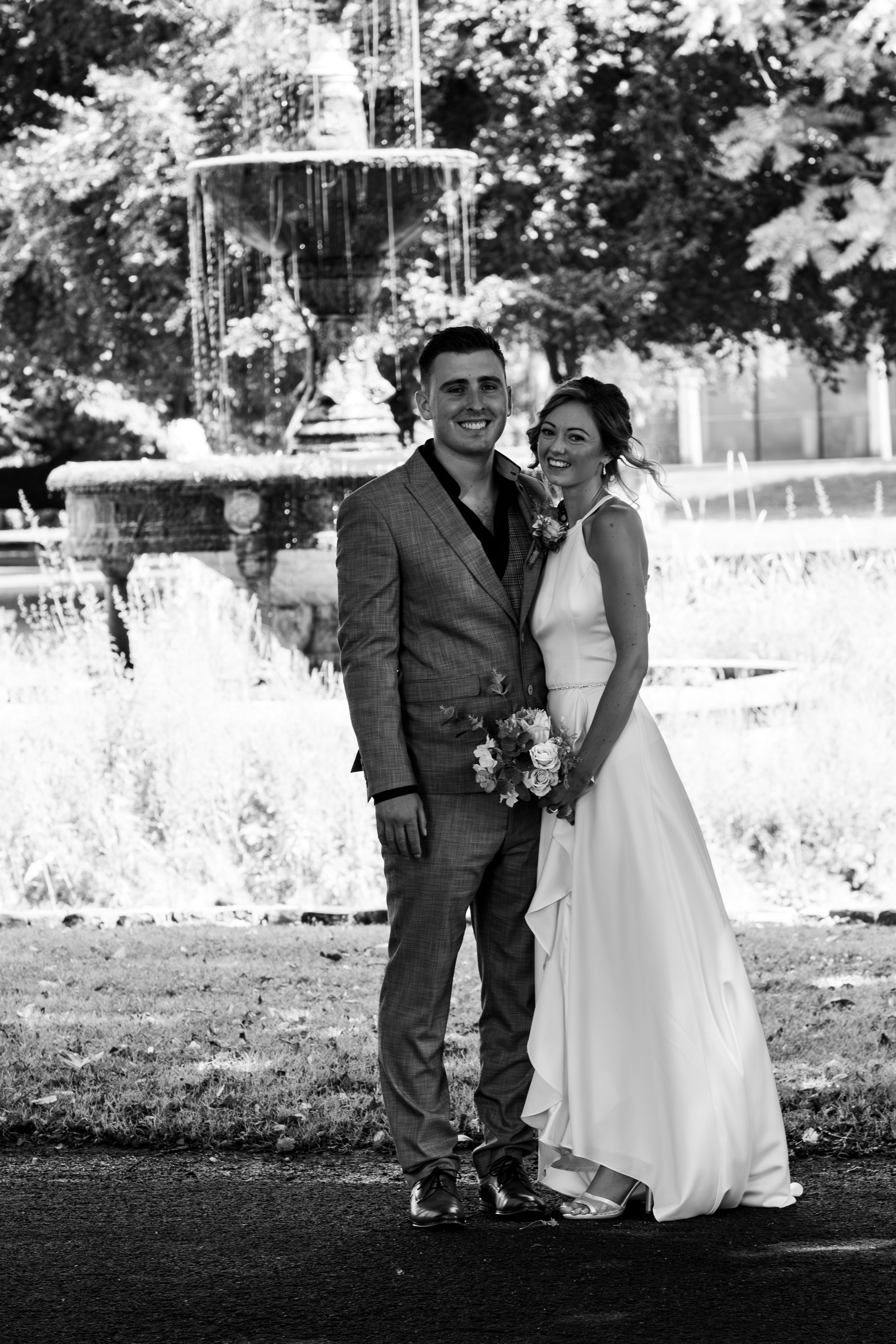 A bride and groom smiling and standing arm in arm outdoors in front of a fountain, with trees in the background. The bride is holding a bouquet and wearing a wedding dress. The groom is dressed in a suit.