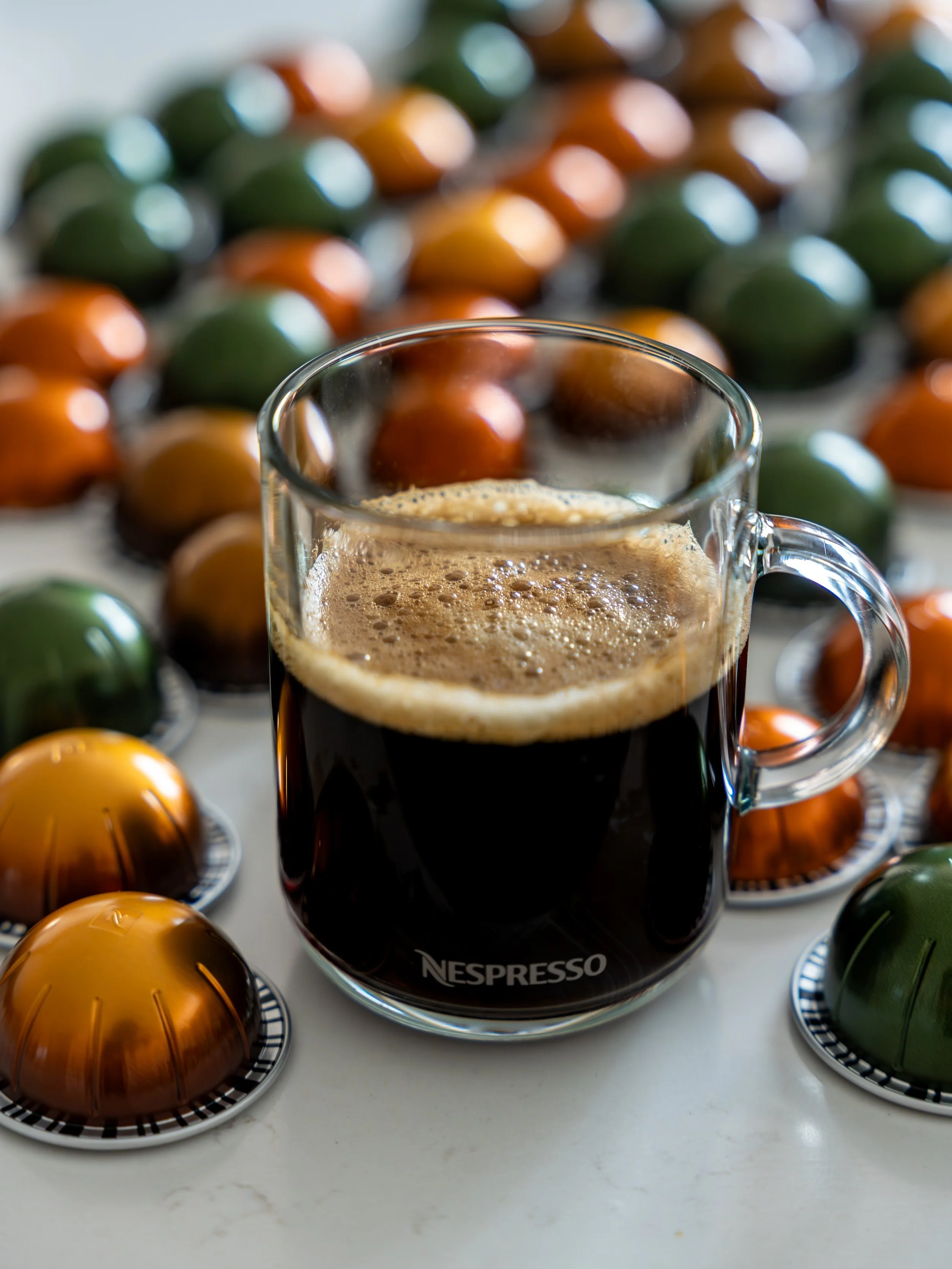A glass mug of black coffee on a white surface, surrounded by colorful tea light candles in green, orange, and gold.