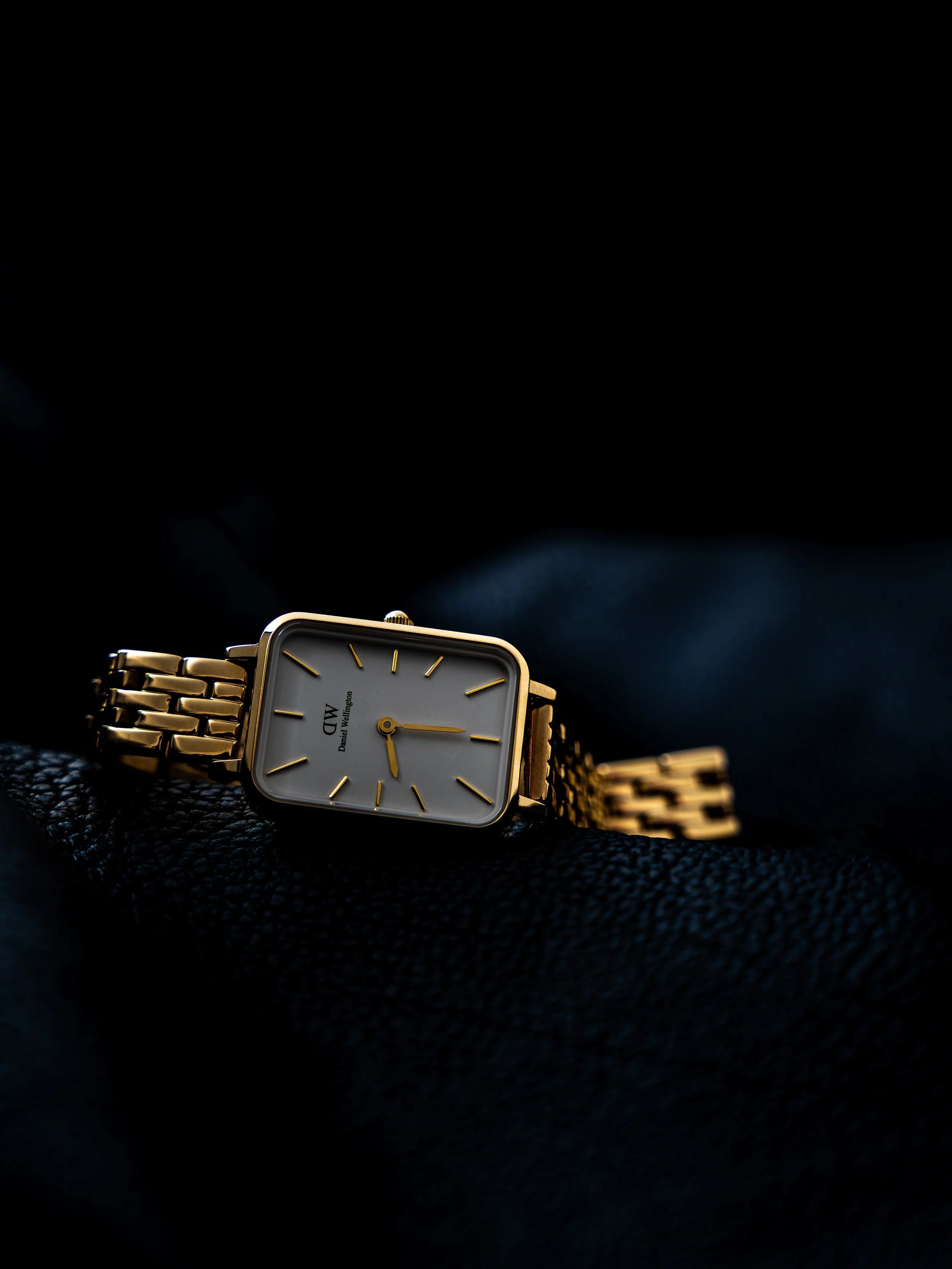 A gold-colored wristwatch with a square white face, gold hour markers, and a gold band resting on black leather fabric.