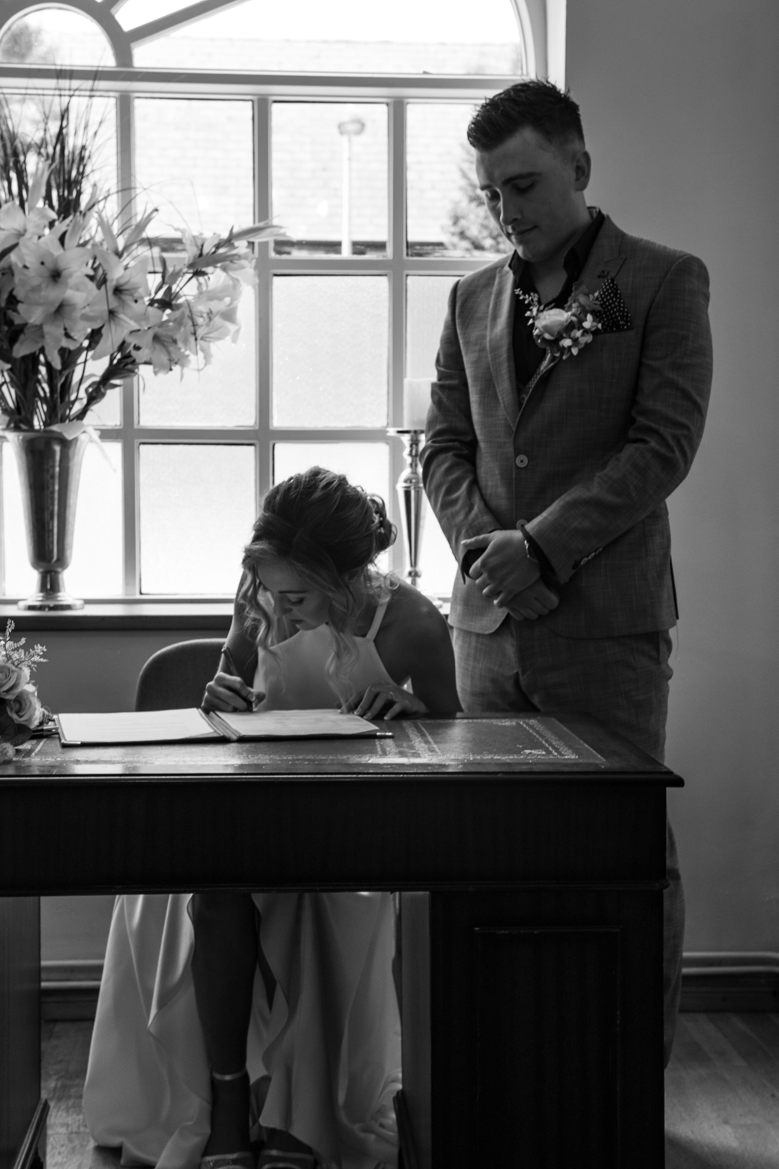 A woman signing a document at a table as a man stands beside her placed in a room with a large window and a flower arrangement.