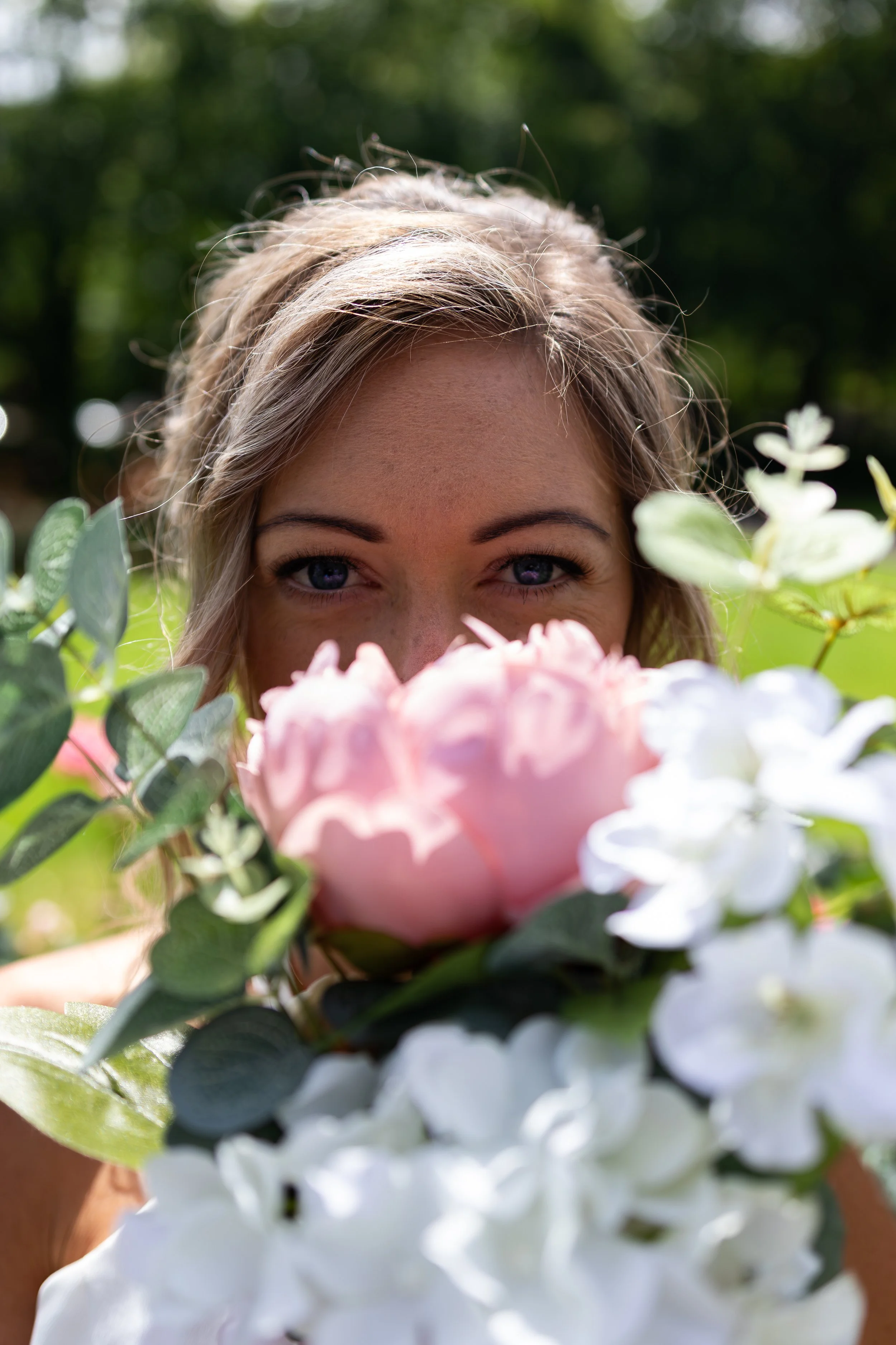 A woman with blonde hair and blue eyes peeking over a bouquet of pink and white flowers with green foliage, outdoors in a sunny setting.