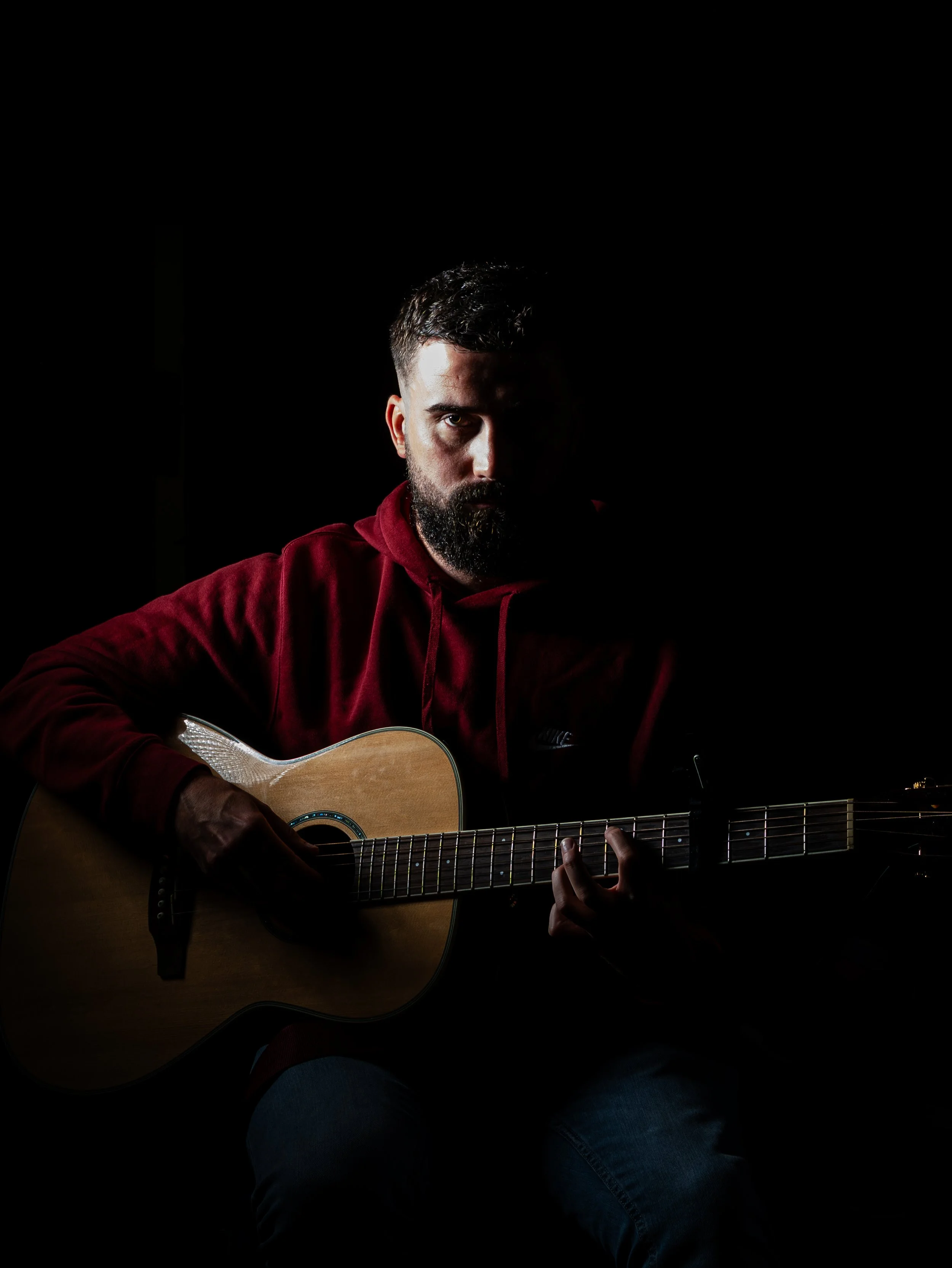 Man with beard wearing red hoodie playing an acoustic guitar against a dark background.