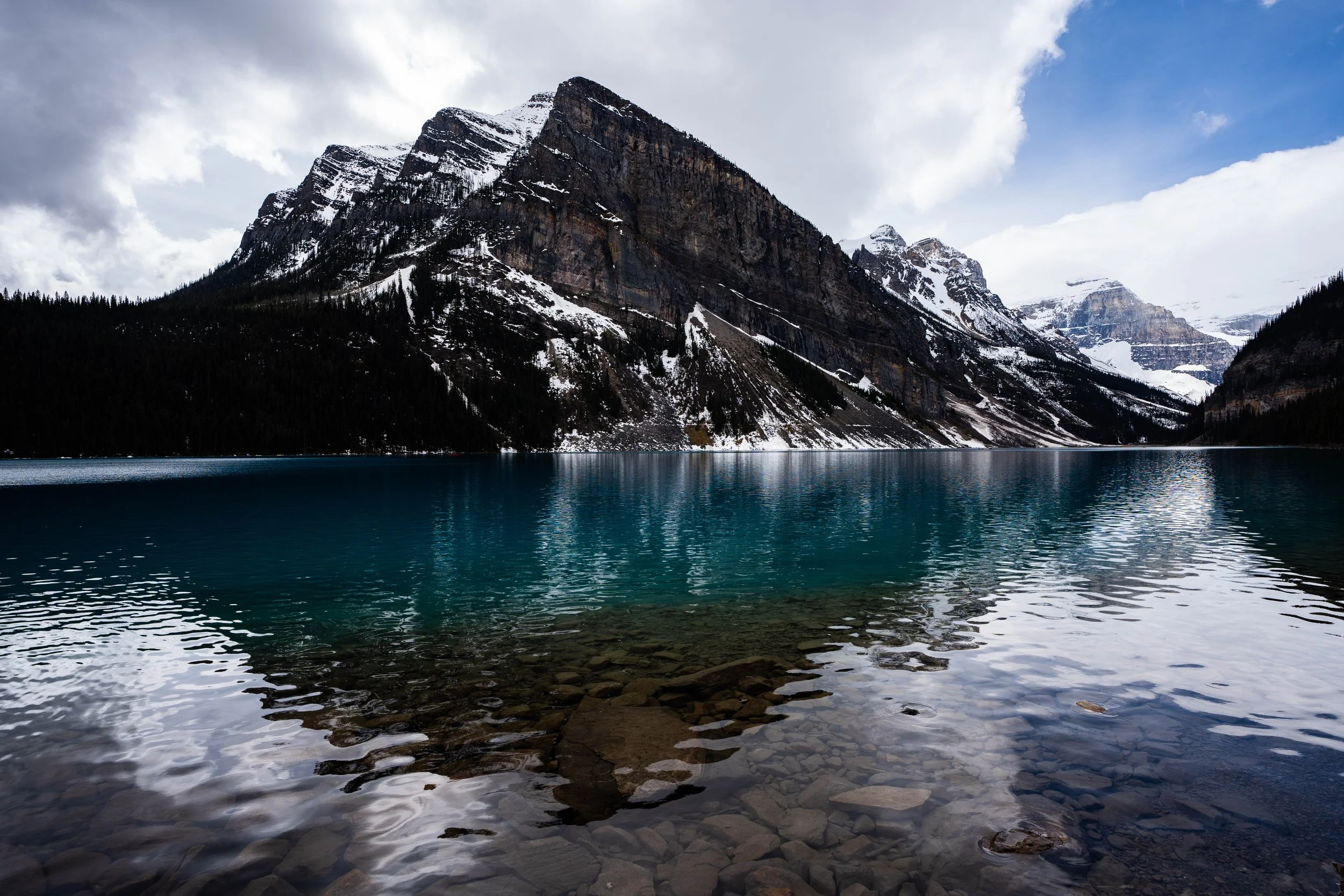 Snow-capped mountains beside a calm blue lake with rocky shoreline and partly cloudy sky.