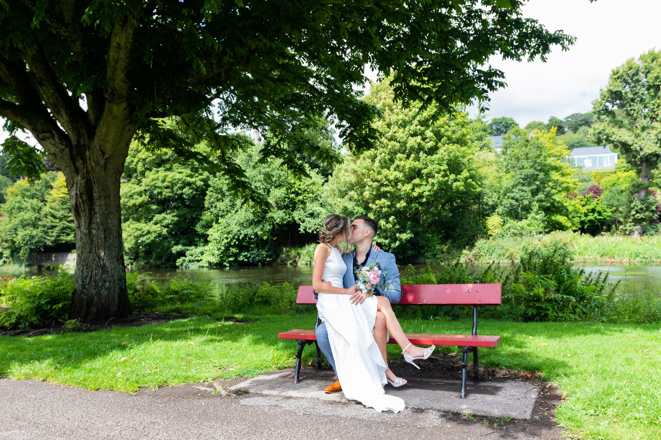 A couple dressed in wedding attire sharing a kiss on a park bench near a river, surrounded by lush green trees and foliage.