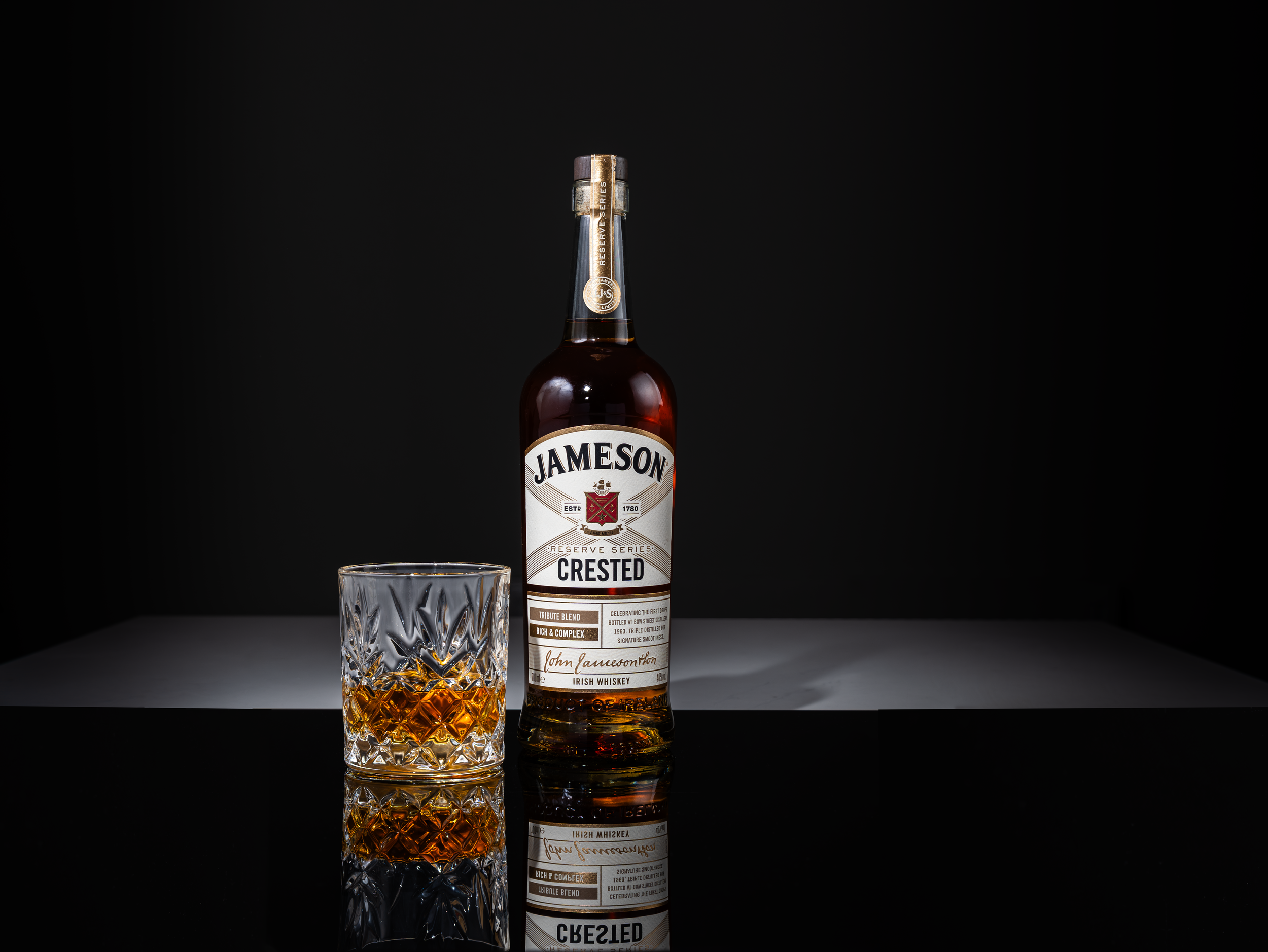 A bottle of Jameson crested Irish whiskey and a glass filled with whiskey on a reflective surface against a black background.