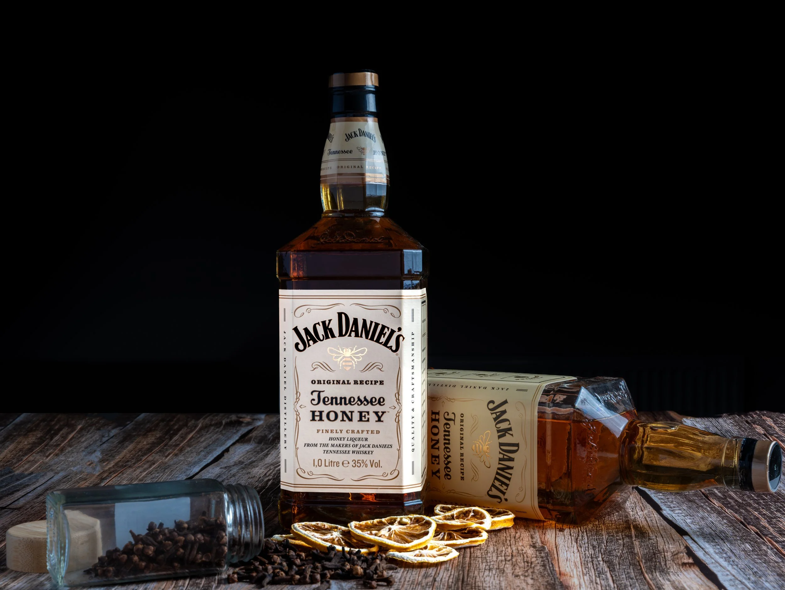 A bottle of Jack Daniel's Tennessee Honey whiskey stands upright on a rustic wooden surface, with its label visible. Another bottle of Jack Daniel's Tennessee Honey is lying on its side nearby. In the foreground, there is a small glass jar tipped ove