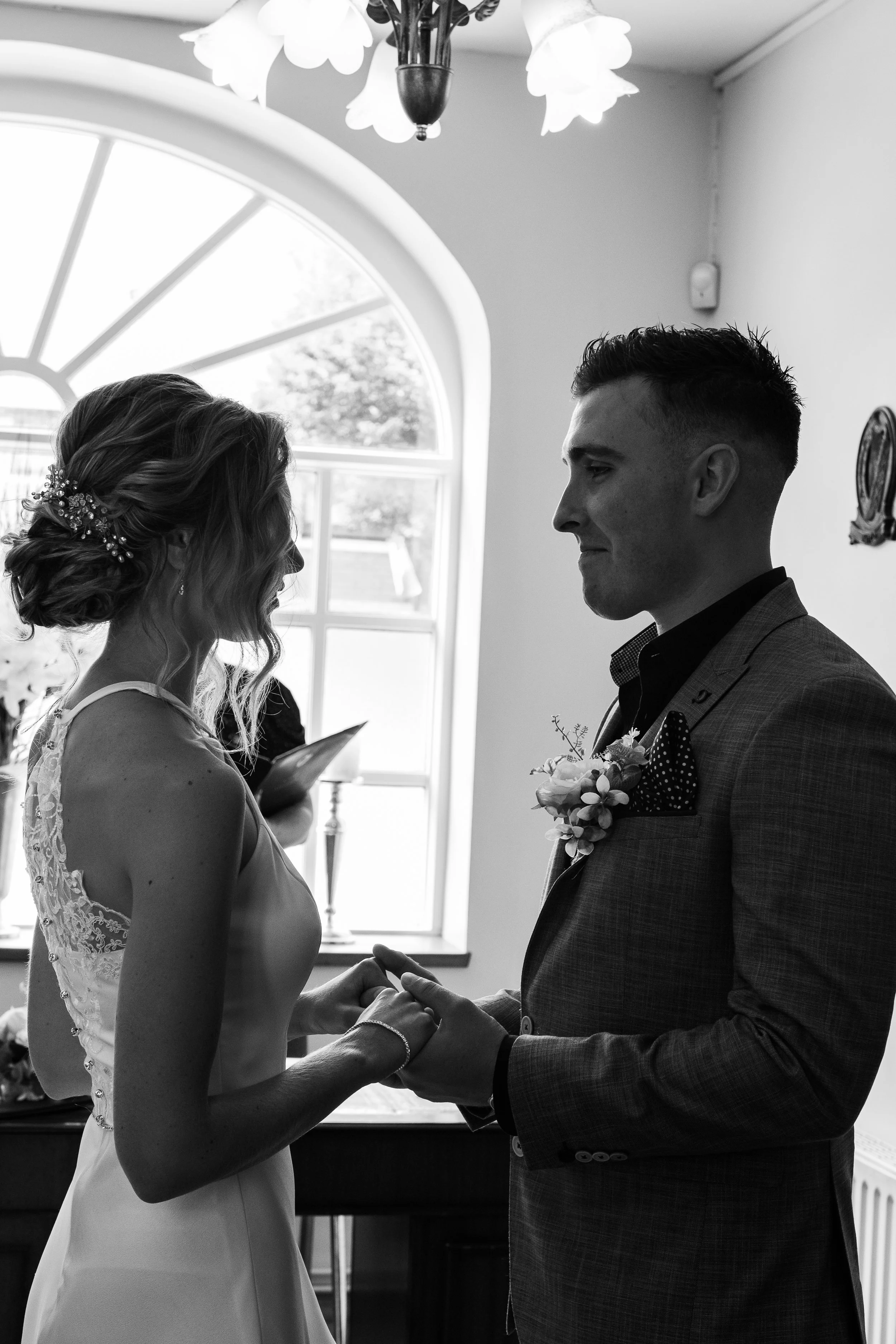 A black and white photo of a bride and groom during their wedding ceremony, holding hands and facing each other indoors near a large window, with a person reading from a book in the background.
