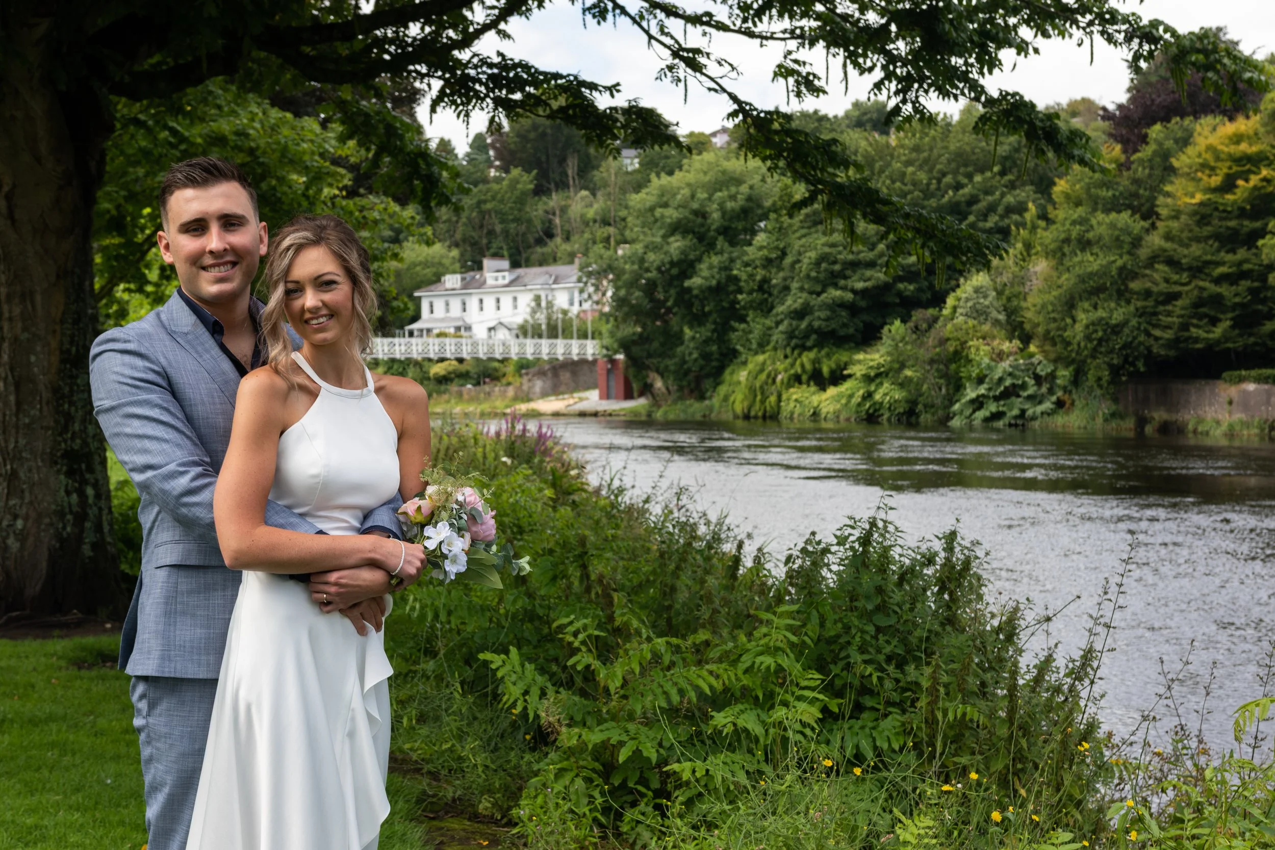 A newlywed couple posing by a river in a lush green park with trees and a white house in the background.