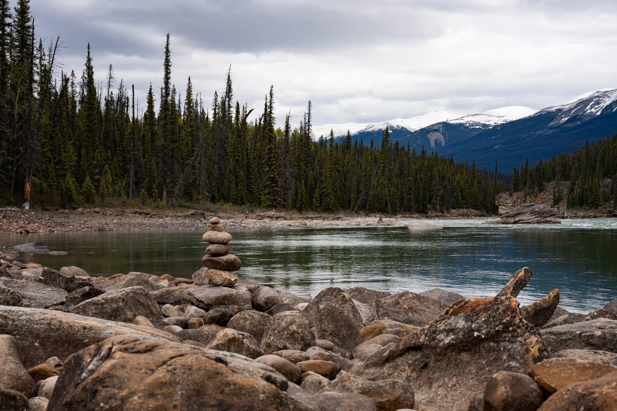 A river flows through a rocky shoreline with a balanced stone stack in the foreground, dense evergreen forest on the opposite bank, snow-capped mountains under a cloudy sky.