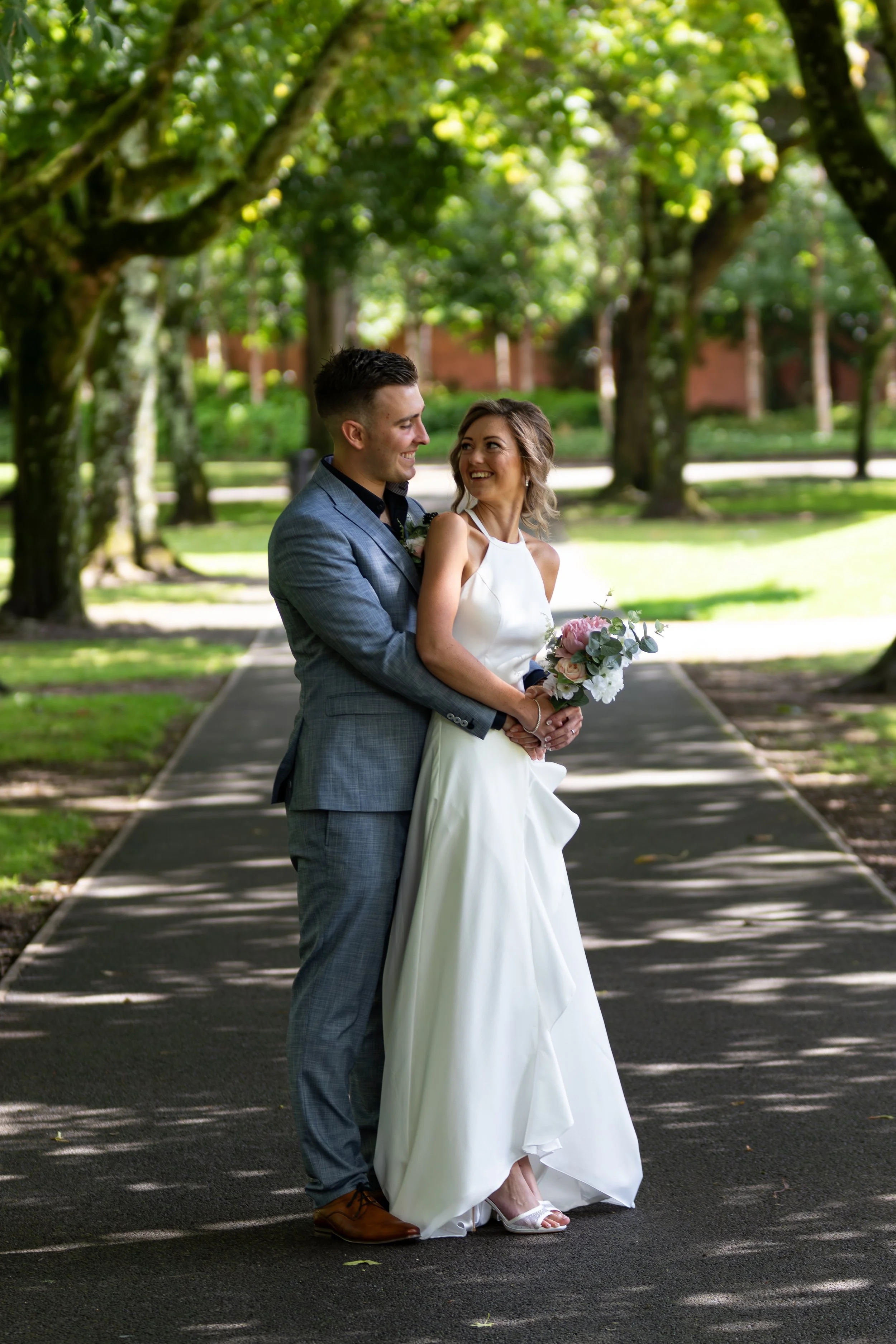 A newlywed couple stands on a shaded park pathway, smiling at each other; the bride holds a bouquet of pink and white flowers, and the groom is dressed in a gray suit.