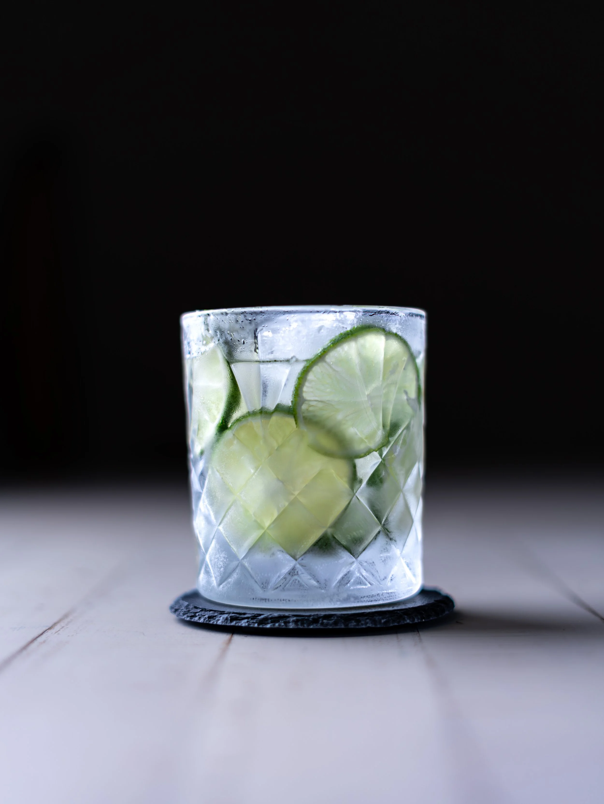 Cork City Product photos ,A clear glass with a diamond pattern filled with ice and slices of lime, placed on a black coaster on a white surface, with a black background.